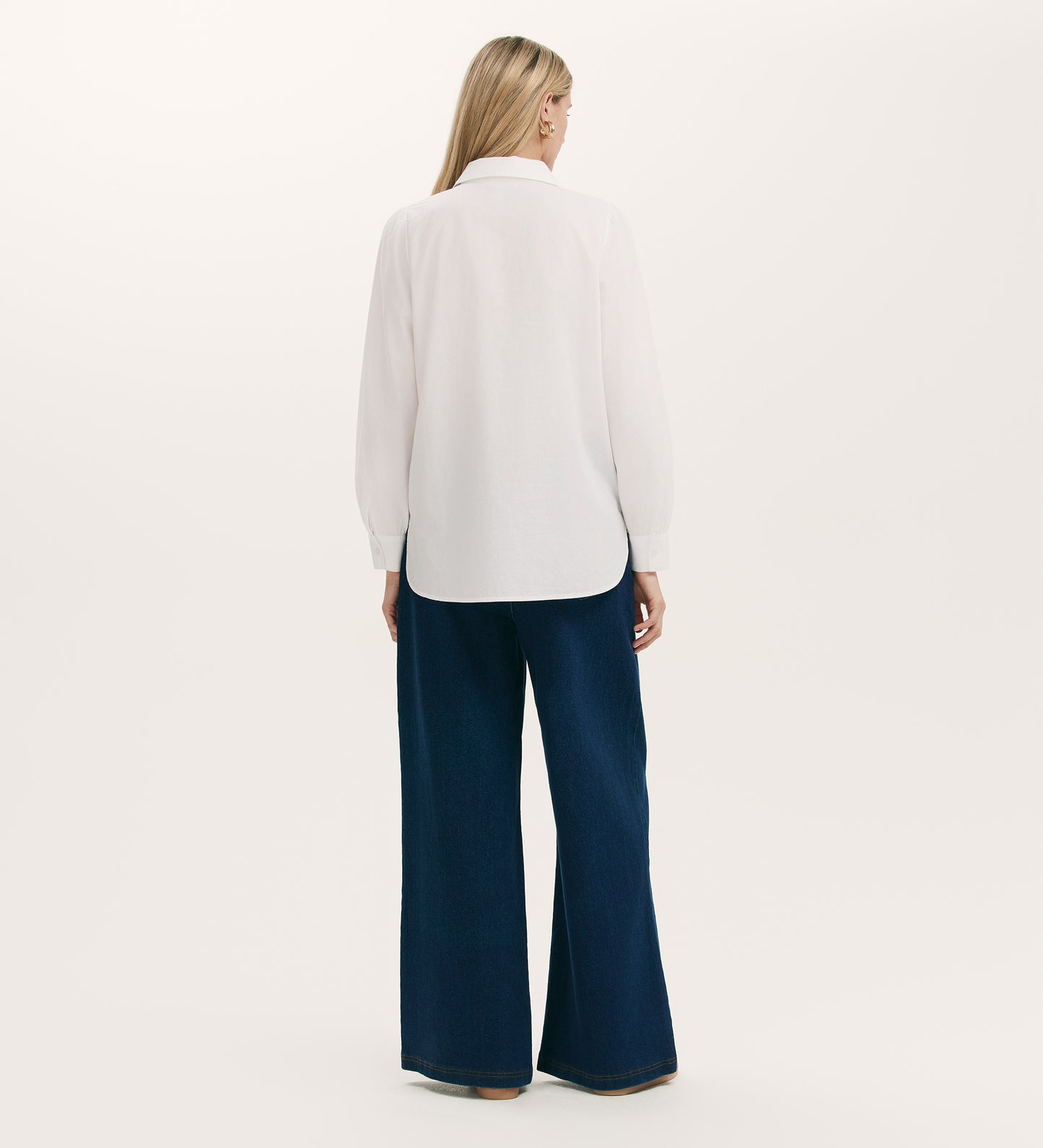 A woman with long blonde hair is seen from the back, wearing the Remy White Cotton Shirt and wide-leg dark blue jeans, standing against a plain light background.