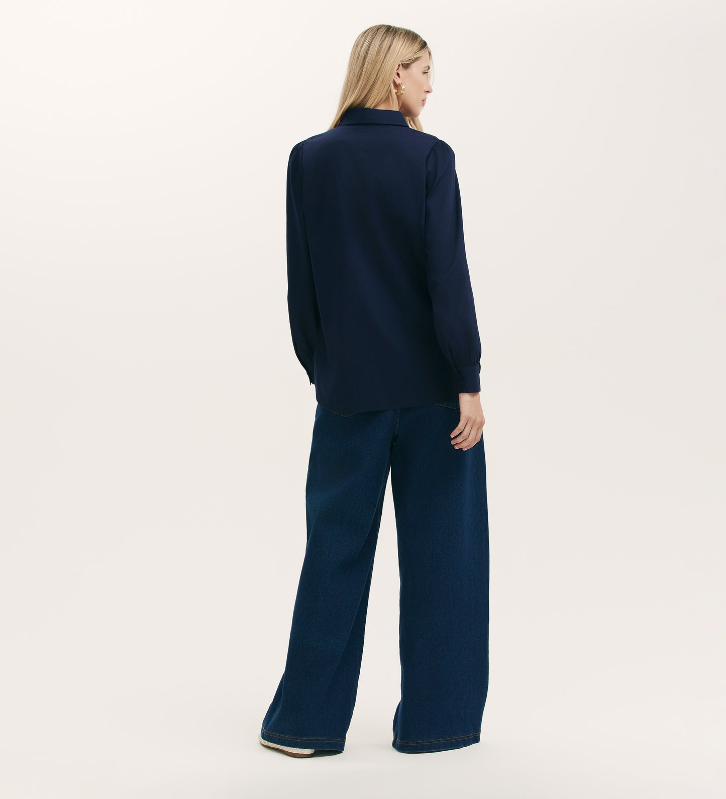 A woman stands facing away, wearing the Remy Navy Cotton Shirt with pintuck accents and wide-leg dark blue jeans against a plain light background.
