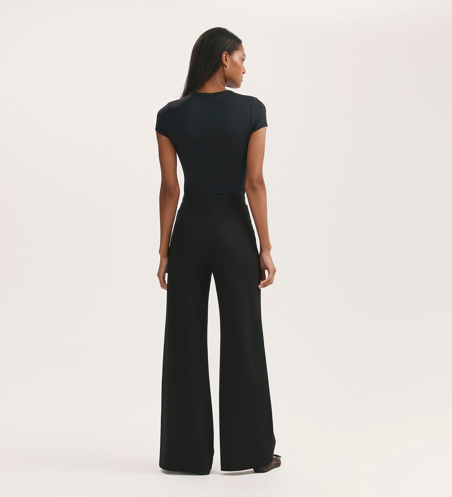 A woman faces away from the camera, wearing a fitted black short-sleeve top and the Reyna Black Ponte Jersey Wide Leg Trousers against a plain, light background.