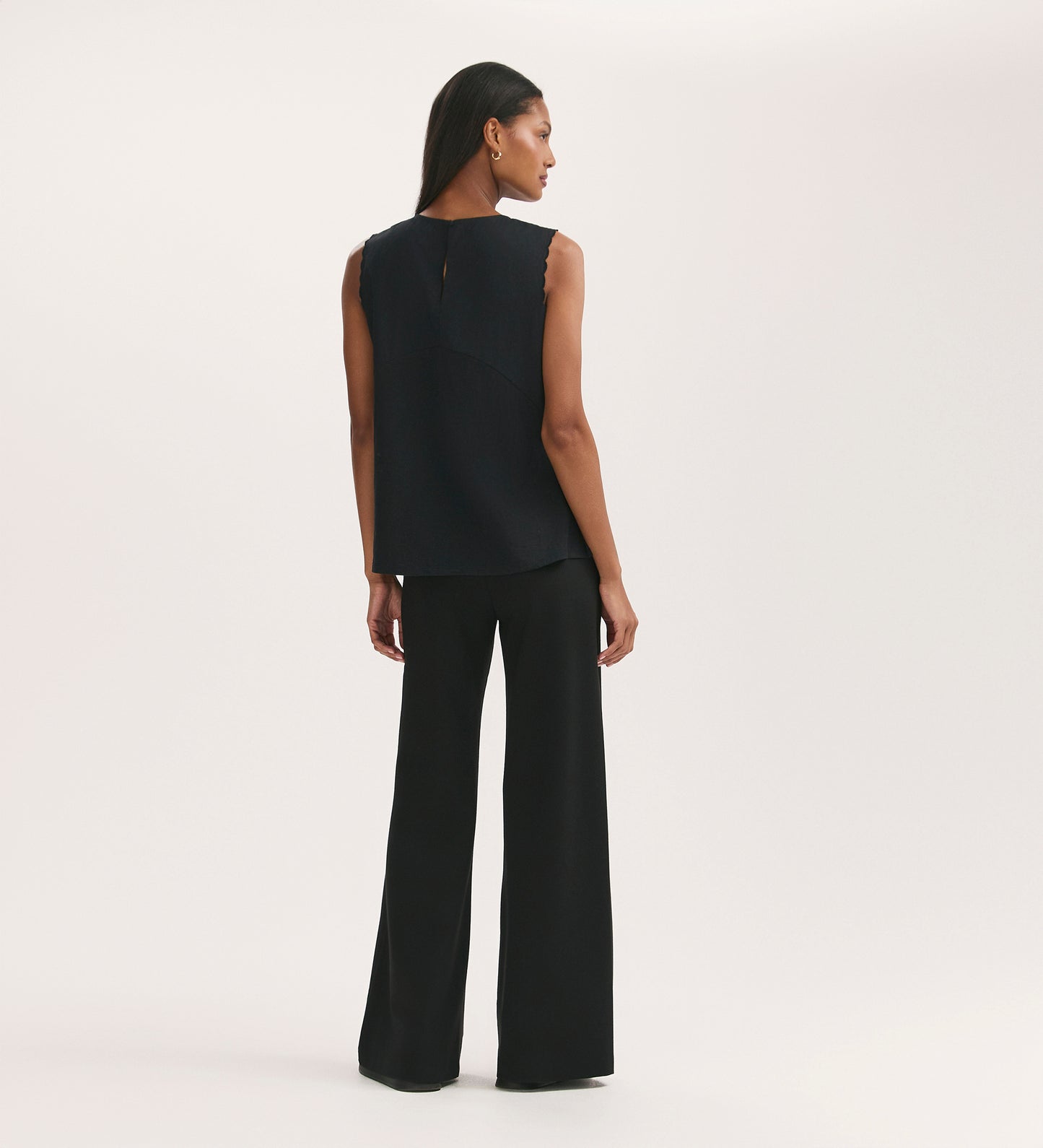 A woman faces away against a plain light background, wearing the Chrissy Black Cotton Sleeveless Top and wide-leg black pants. Her straight hair is tucked behind her ears, and one arm rests relaxed at her side.