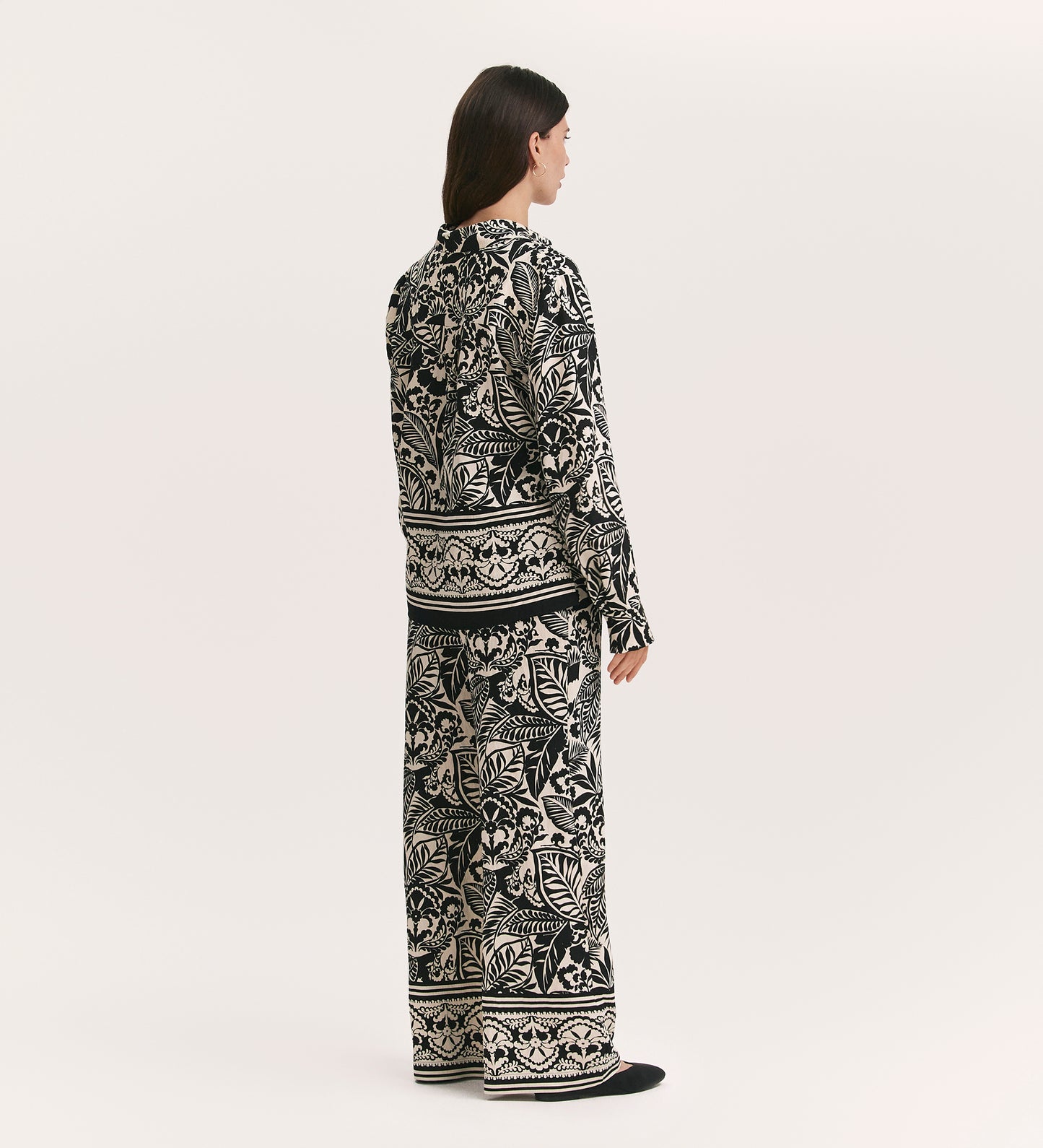 A person with long dark hair, seen from behind, wears the Roma Black Border Leaf Crepe Button Through Shirt with wide-leg pants, presenting a sophisticated look against a plain light background.