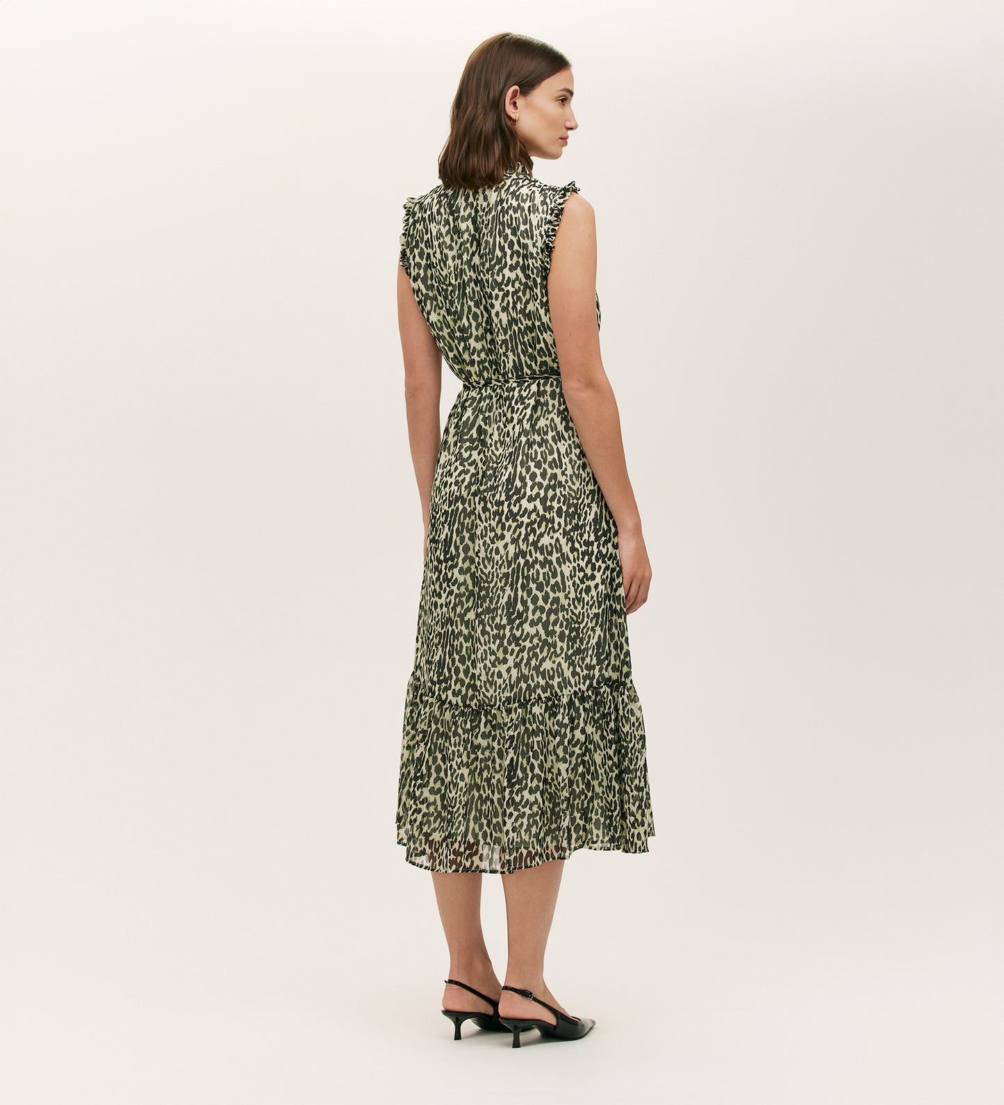 A woman with shoulder-length brown hair models the Rosalind Green Leopard Chiffon Sleeveless Midi Dress, featuring ruffled tiers and an animal print, paired with black heeled sandals, standing against a plain white background, facing away.