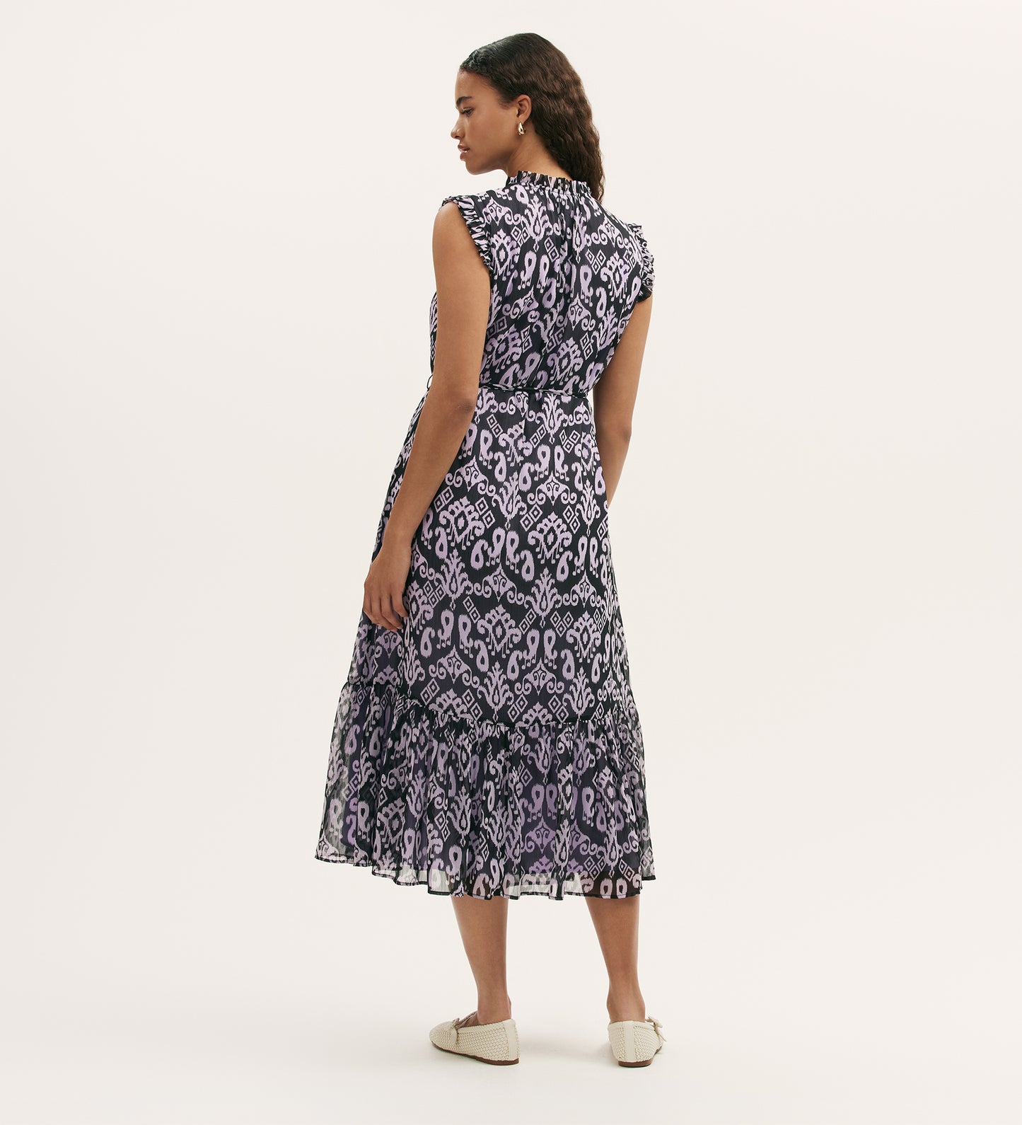 A woman stands facing away, wearing the Rosalind Lilac Ikat Chiffon Sleeveless Midi Dress with a tiered skirt and white flats against a plain, light background.