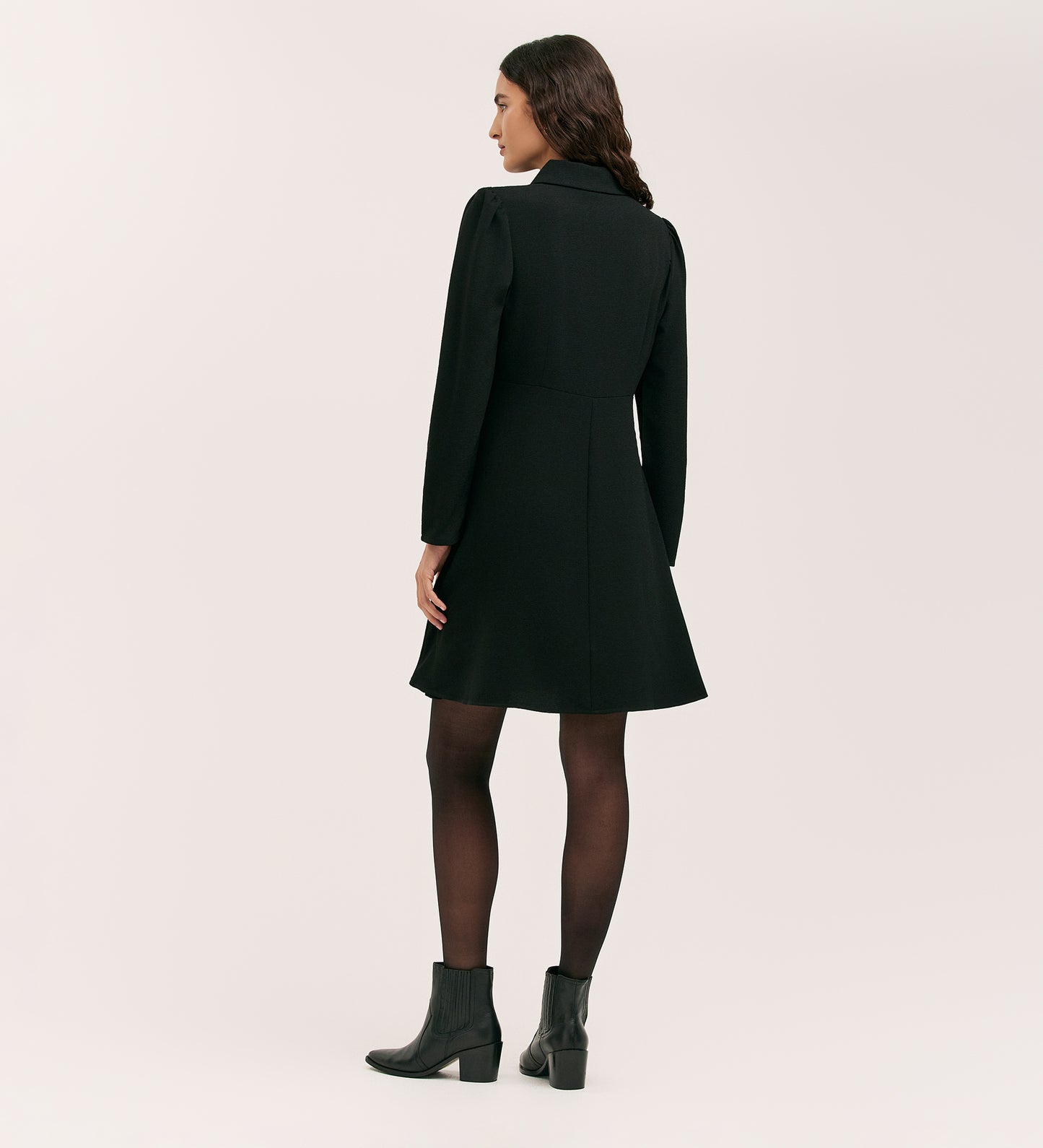 A woman with long wavy brown hair, seen from behind, wears the Sabine Black Crepe Short Dress styled with black tights and ankle boots, set against a plain light background.