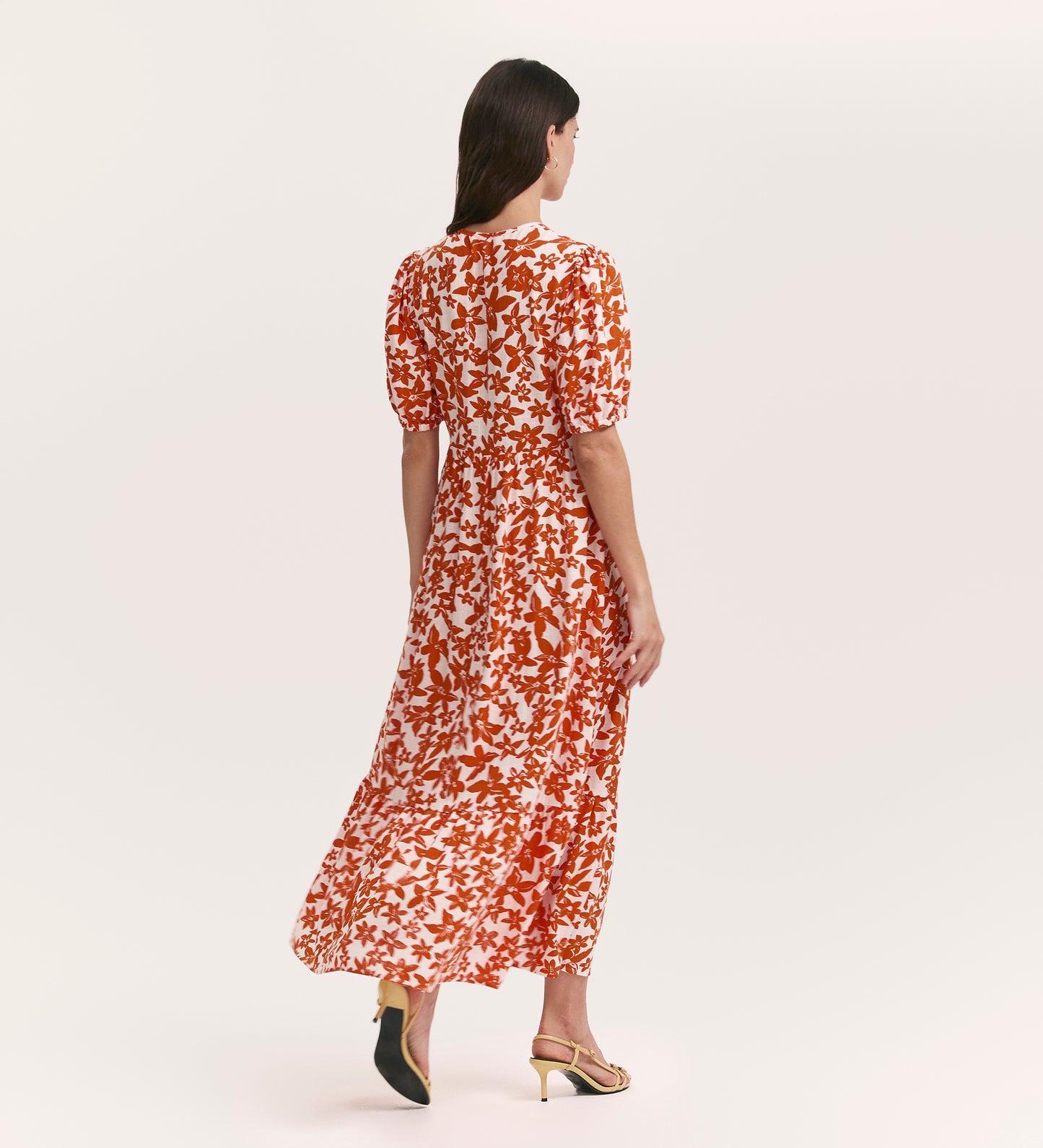 A woman with long brown hair stands facing away in the Sabine Orange Flowers Midi Tiered Dress, featuring a v-neckline and short sleeves, paired with open-toed sandals against a plain light background.