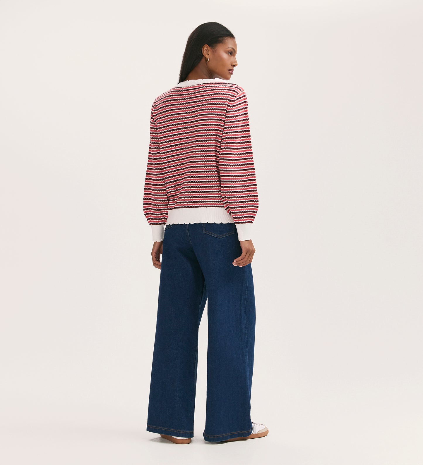 A woman with straight dark hair models the Sanne Pink Stripe Jumper, a relaxed fit knitwear sweater in pink stripes with scalloped edges. She pairs it with wide-leg blue jeans and beige shoes, standing against a plain light background.