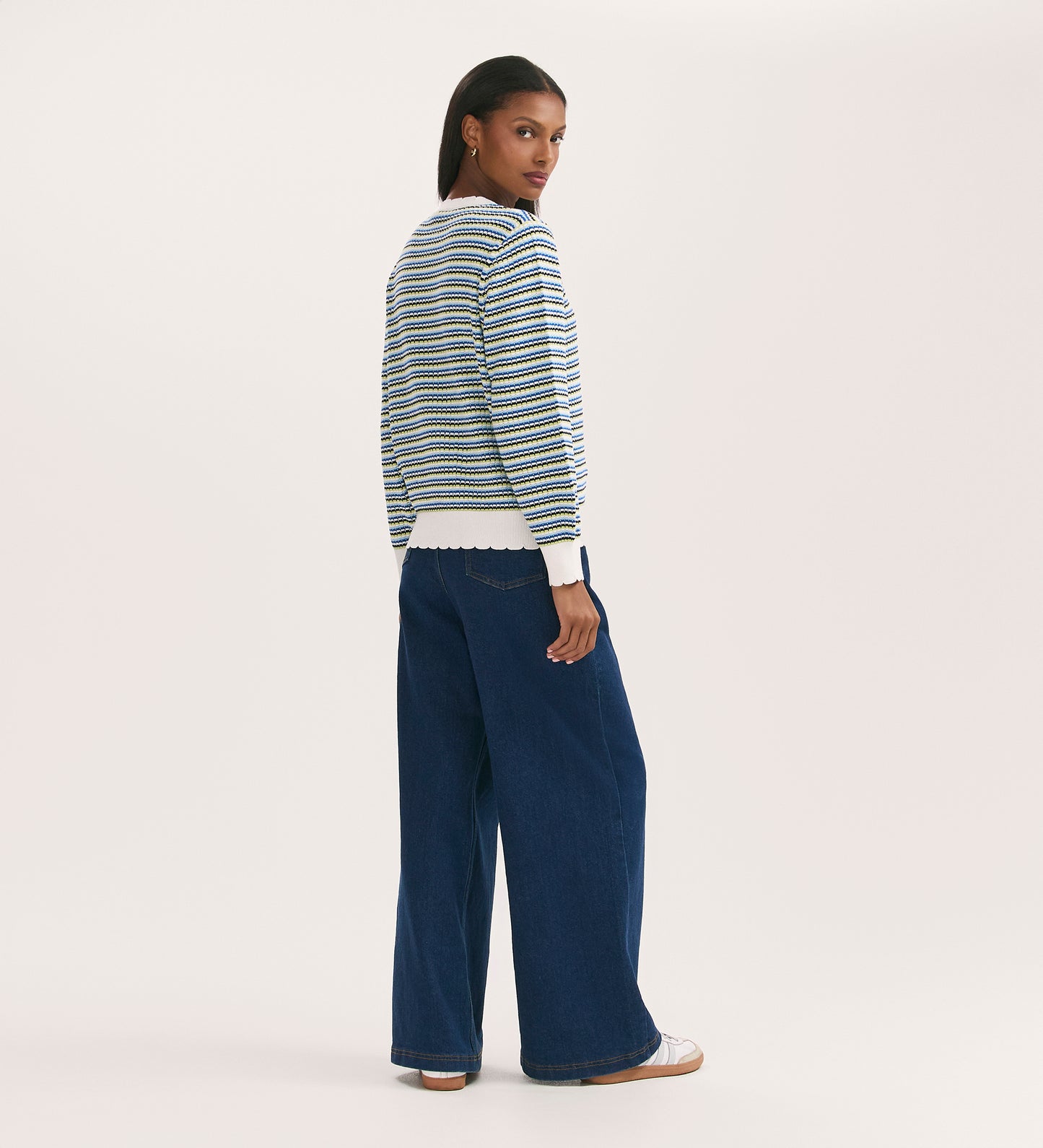 A woman looks over her shoulder, wearing the Sanne Blue Stripe Jumper with wide-leg blue jeans and white sneakers, set against a plain light background.