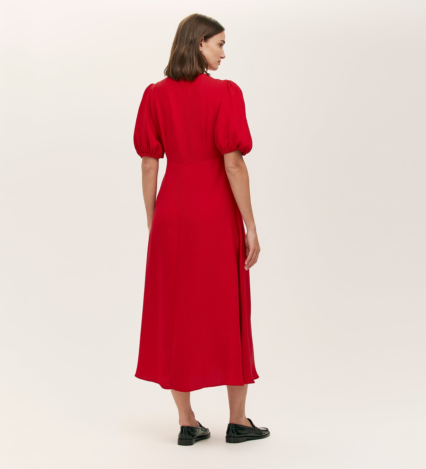 A woman with straight, shoulder-length brown hair wears the Sarah Red Crepe Button Through Midi Tea Dress, featuring gold buttons and short puff sleeves. She faces away in black shoes against a plain, light-colored background.