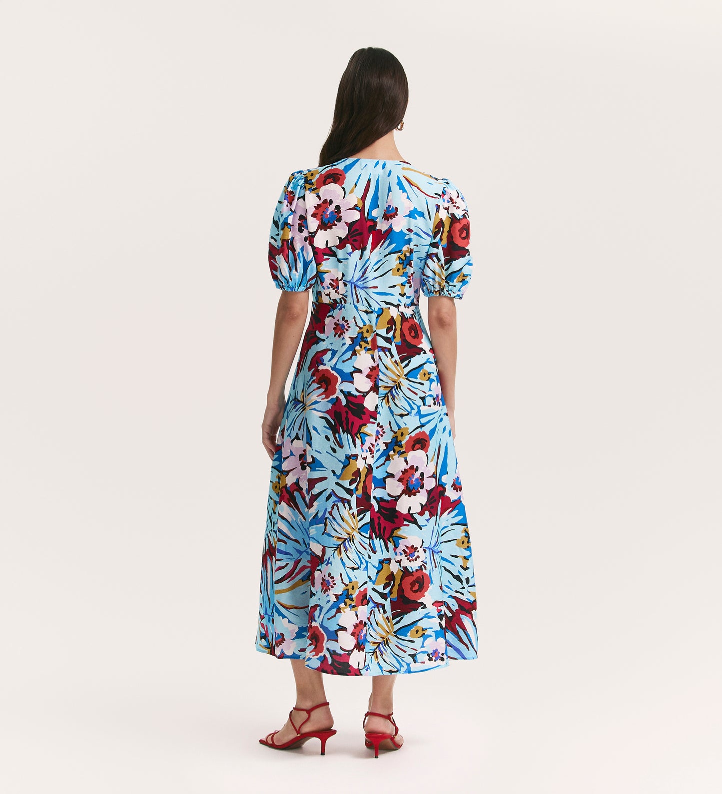 A woman with long brown hair wears the Sarah Aqua Floral Crepe Midi Dress, featuring bold colorful floral prints and a flattering v-neckline. She pairs it with red strappy heels while standing against a plain white background.