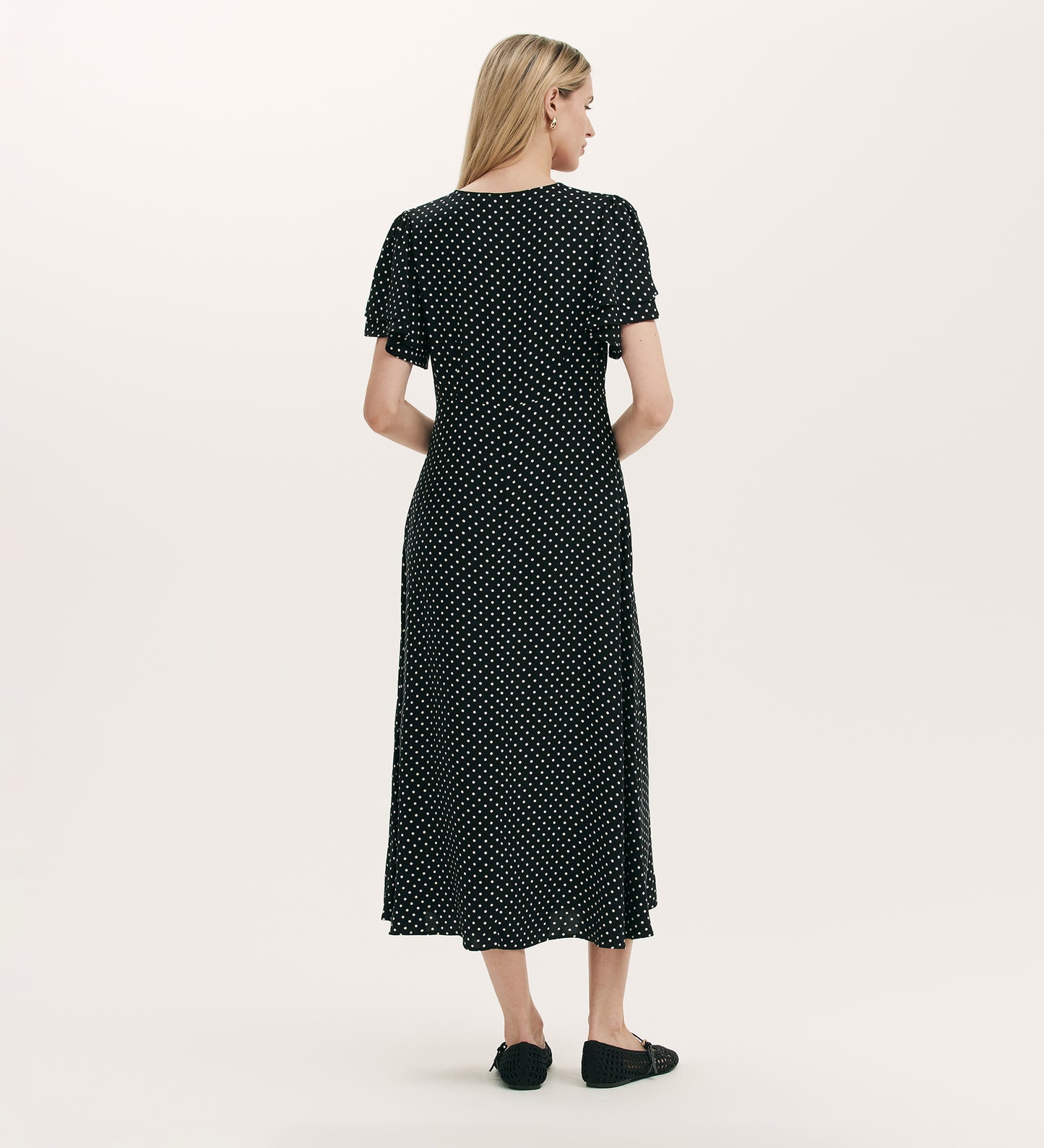 A woman stands with her back to the camera, wearing the Saskia Black Polka Dot Midi Tea Dress featuring angel sleeves and an empire waist. She pairs it with black flats against a plain light beige background.