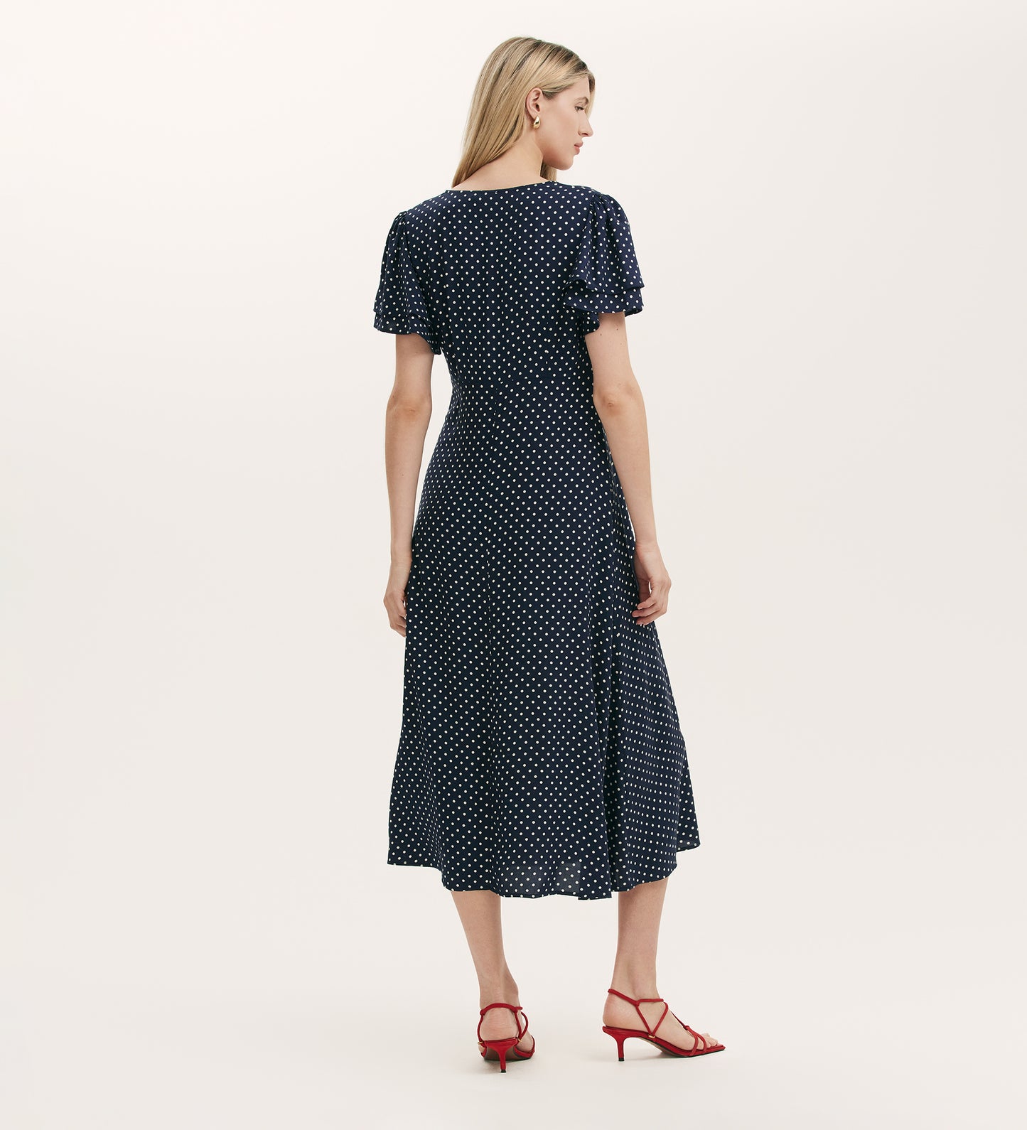 A woman with long blonde hair, facing away, wears the Saskia Navy Polka Dot Midi Tea Dress and red heeled sandals against a plain light background.