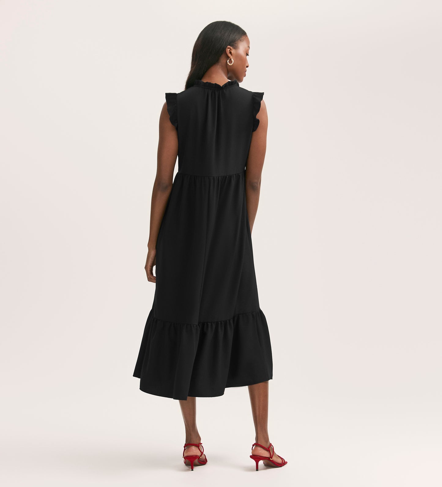 A woman faces away, wearing the Scout Black Crepe Midi Dress—relaxed fit, sleeveless, with ruffled shoulders and hem in crease-free crepe—styled with red heeled sandals against a plain light background.