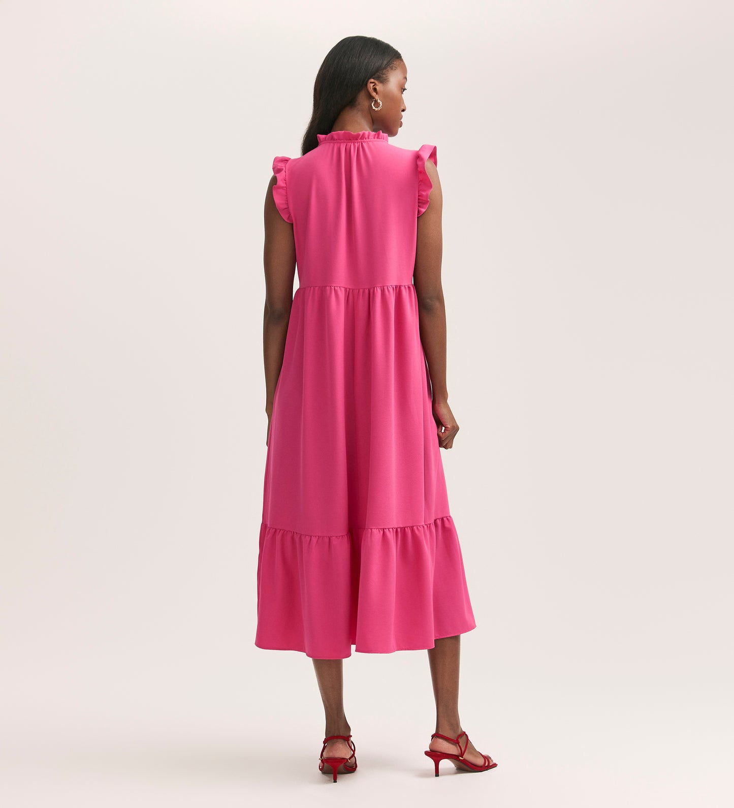 A woman faces away, wearing the Scout Fuchsia Pink Crepe Midi Dress—a relaxed-fit, tiered, sleeveless midi in crease-free crepe—paired with red heeled sandals against a plain light background.