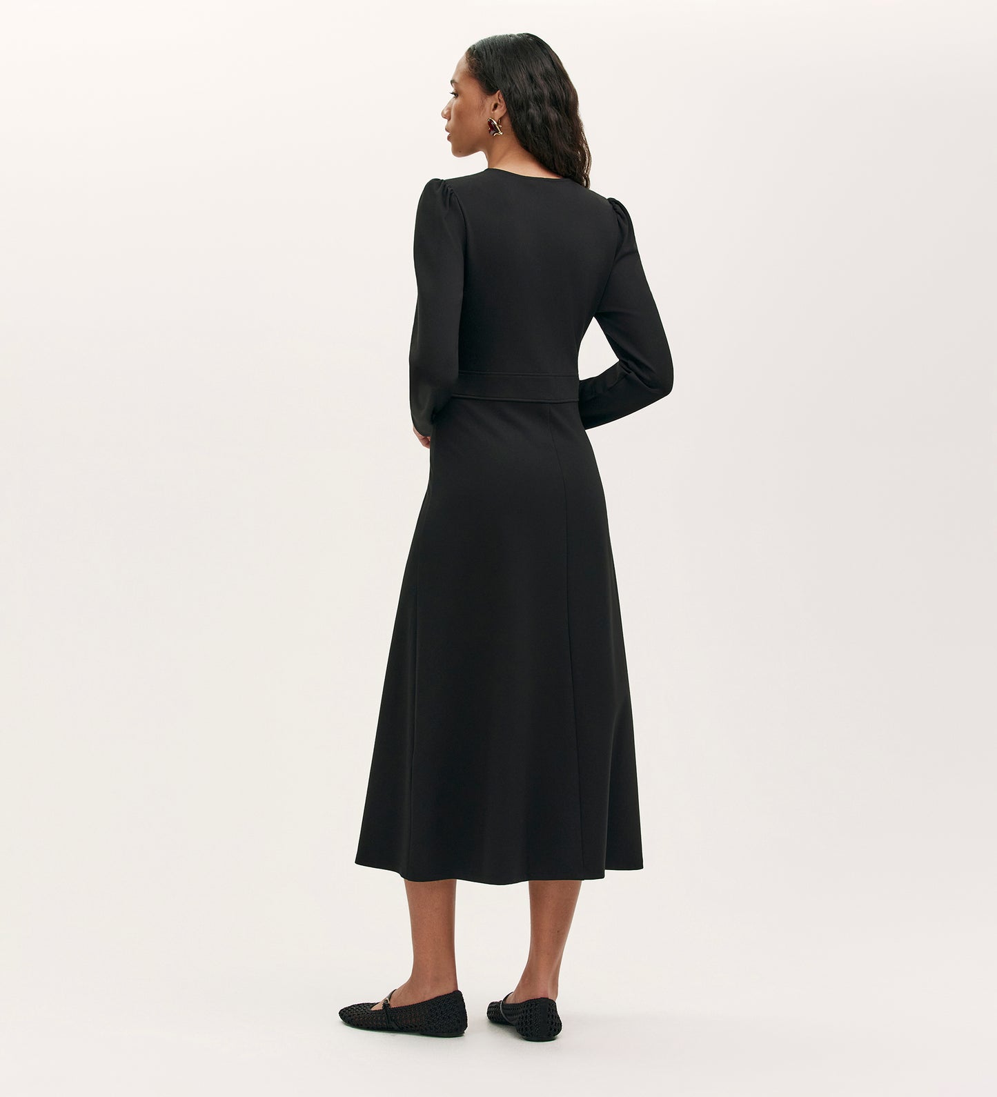 A woman faces away, wearing the Seraphina Black Ponte Jersey Long Sleeve Midi Dress with a fitted waist and black flats, set against a plain light background.