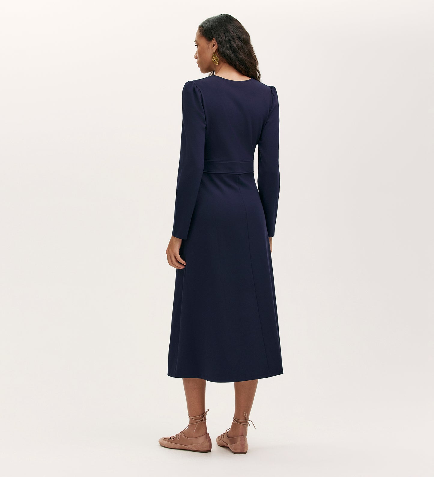 A woman faces away from the camera, wearing the Seraphina Navy Ponte Jersey Long Sleeve Midi Dress with light brown lace-up flats. She completes her look with wavy hair and hoop earrings against a plain, light background.
