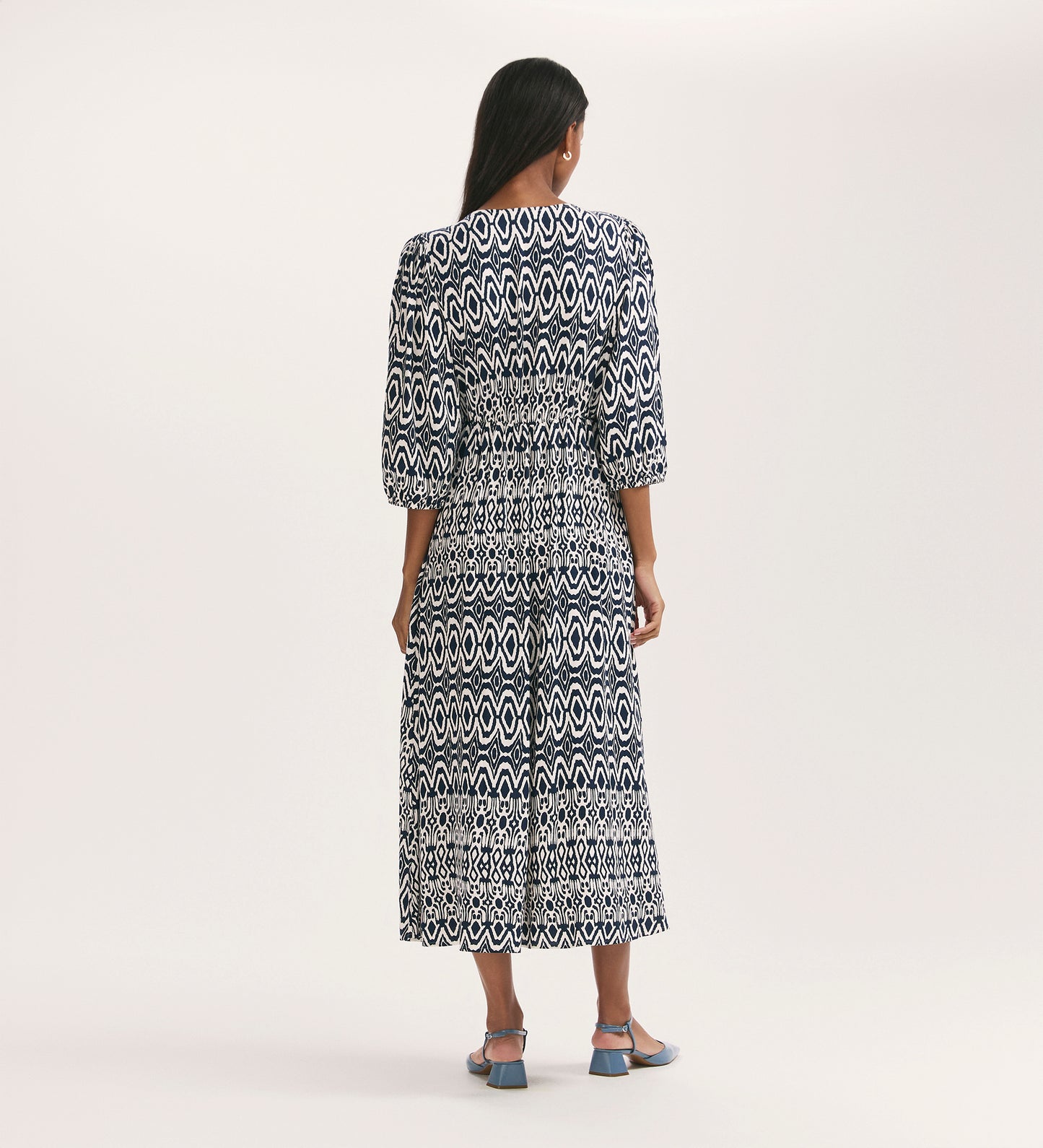 A woman stands facing away in the Serena Navy Tribal Crepe Midi Smock Dress, featuring a fit and flare silhouette with three-quarter sleeves, a cinched waist, and blue heeled sandals, set in a plain light-colored studio.