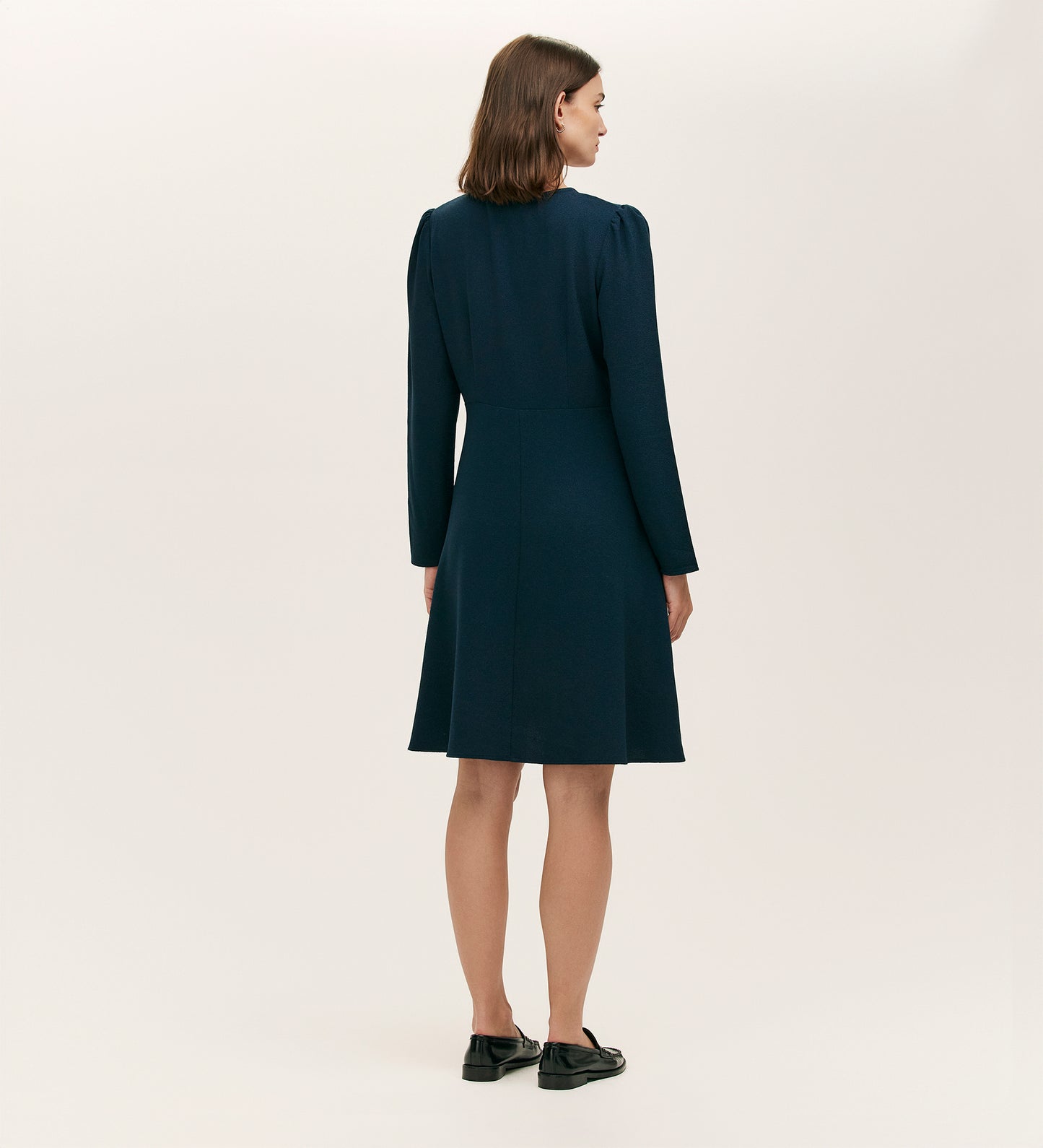 A woman with shoulder-length brown hair faces away, wearing the Shauna Navy Crepe Short Dress featuring gold button details and black shoes against a plain light background.