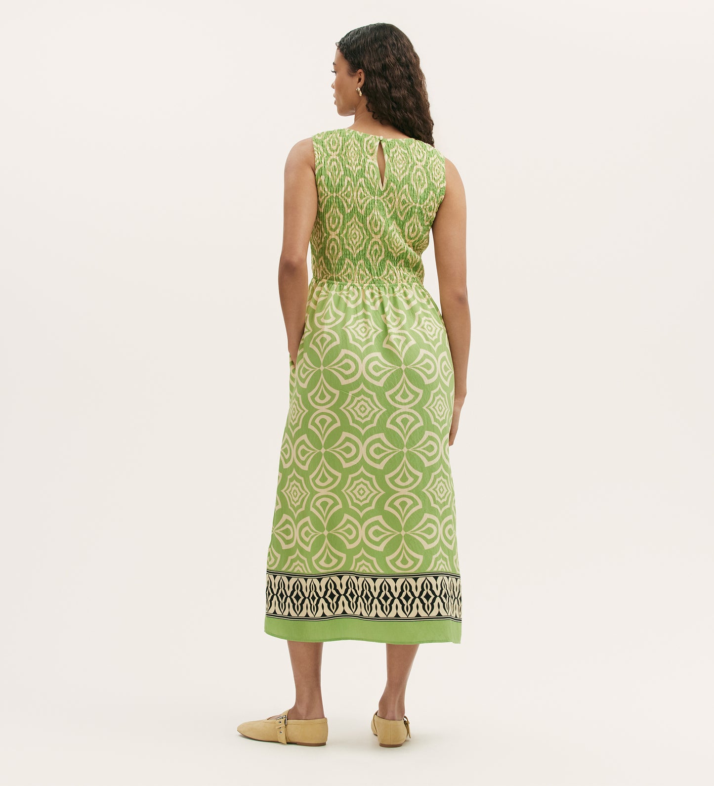 A woman with long, wavy hair wears the Skylar Lime Mosaic Midi Dress featuring a sleeveless design, green mosaic print, keyhole back, and black-and-white border at the hem. She pairs it with beige shoes against a plain light background.