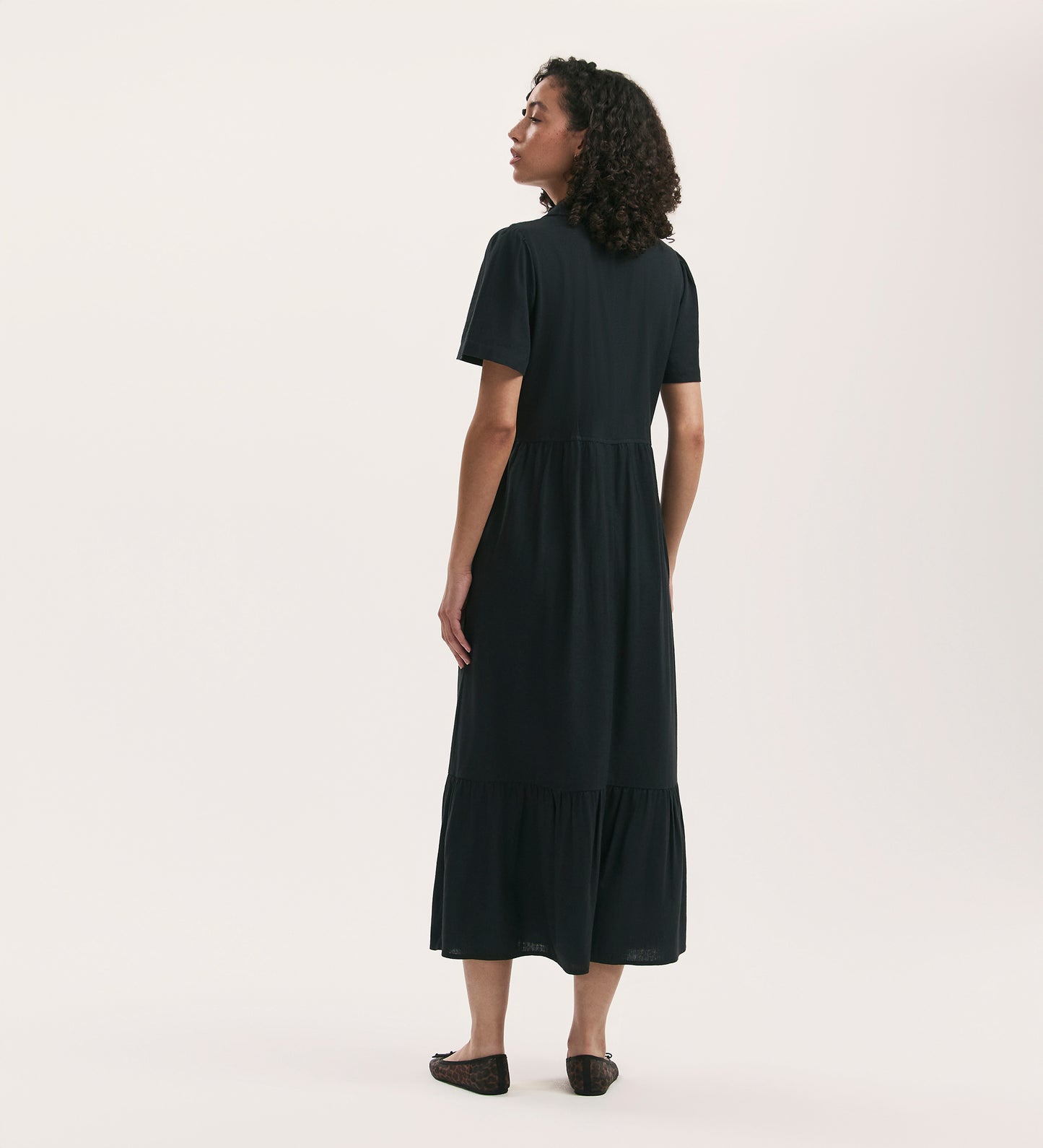 A woman with curly hair faces away, wearing the Tania Black Linen Blend Midi Shirt Dress—relaxed fit, short sleeves, ankle length—paired with flat shoes against a plain, light background.