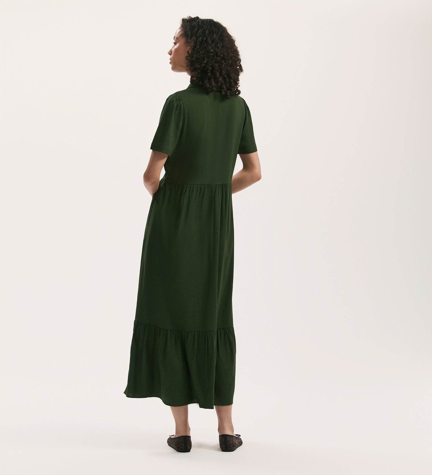 A woman with curly hair stands facing away, wearing the Tania Dark Khaki Linen Blend Midi Shirt Dress in a relaxed fit and black flats against a plain light background.