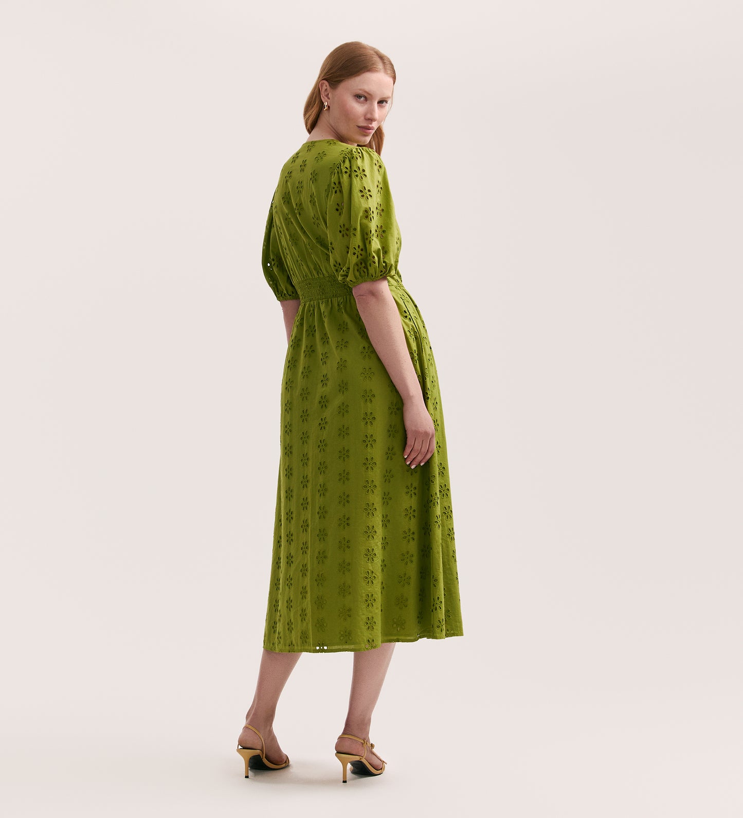 A woman poses in the Una Embroidered Cotton Light Green Midi Tea Dress, featuring short puff sleeves and a subtle floral design. She stands sideways in heeled sandals, looking over her shoulder against a plain light background.