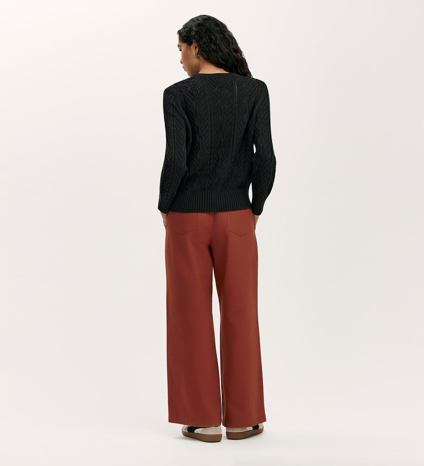 A woman faces away, wearing the Valerie Black Knitted Jumper with a relaxed fit, paired with rust wide-leg pants and brown shoes, set against a plain light background.