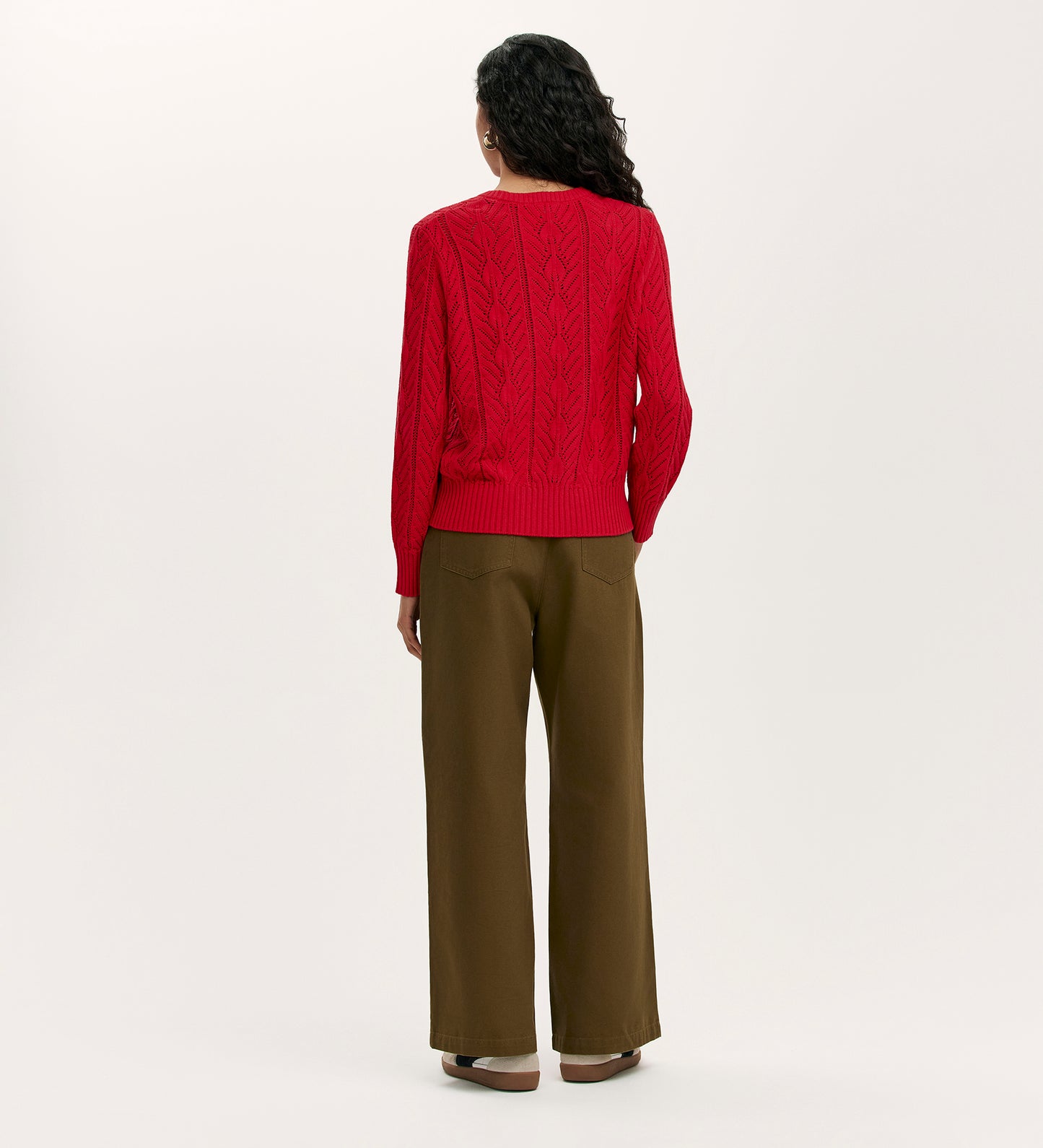 A person with long dark hair faces away, wearing the Valerie Red Knitted Jumper with olive green wide-leg pants and brown shoes against a plain light background.