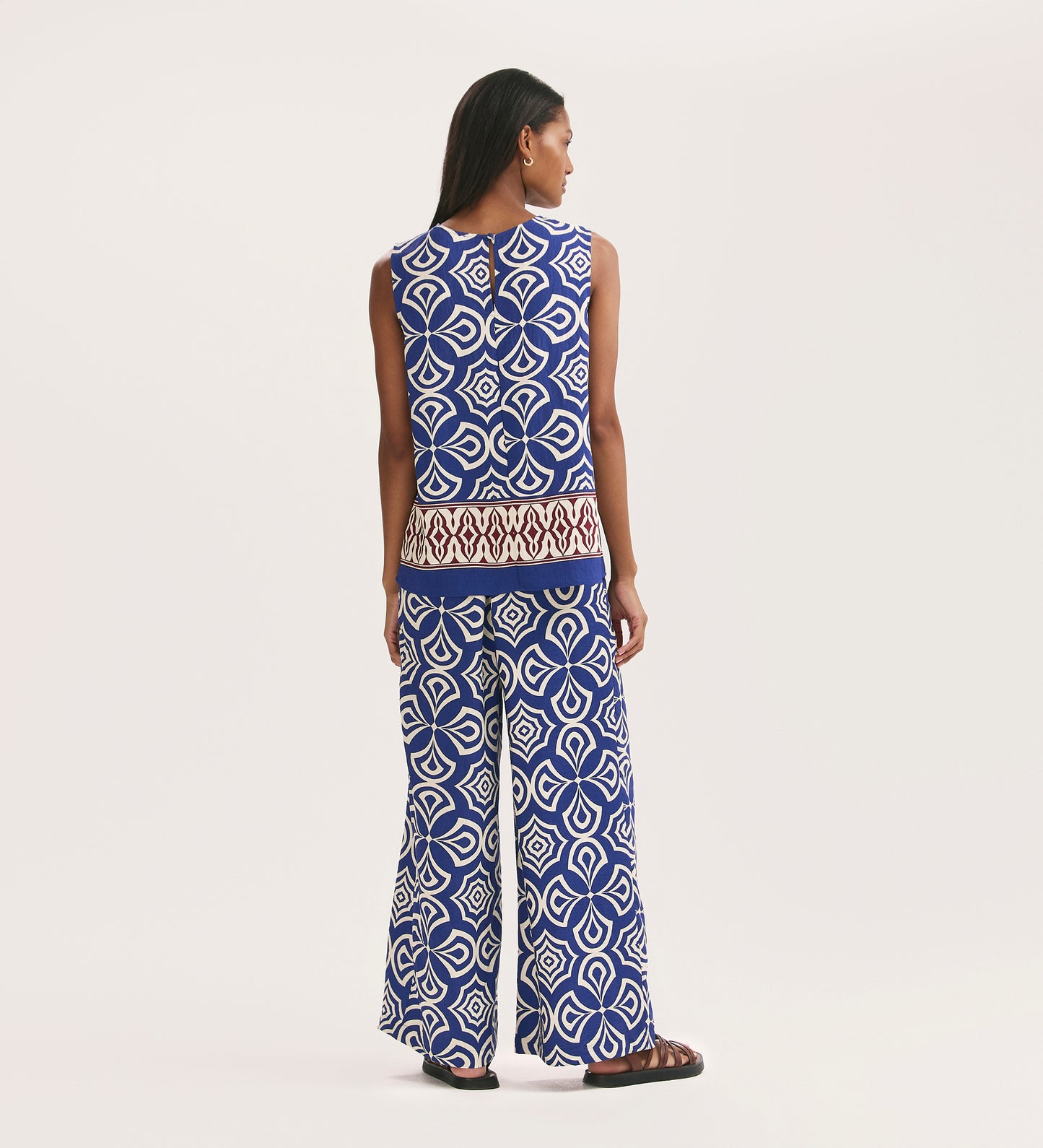 A woman with long hair, in black sandals, faces away wearing Nat Blue Mosaic Crepe Wide Leg Trousers with an elasticated waist and matching sleeveless blue mosaic print top with a brown and white border, set against a plain background.