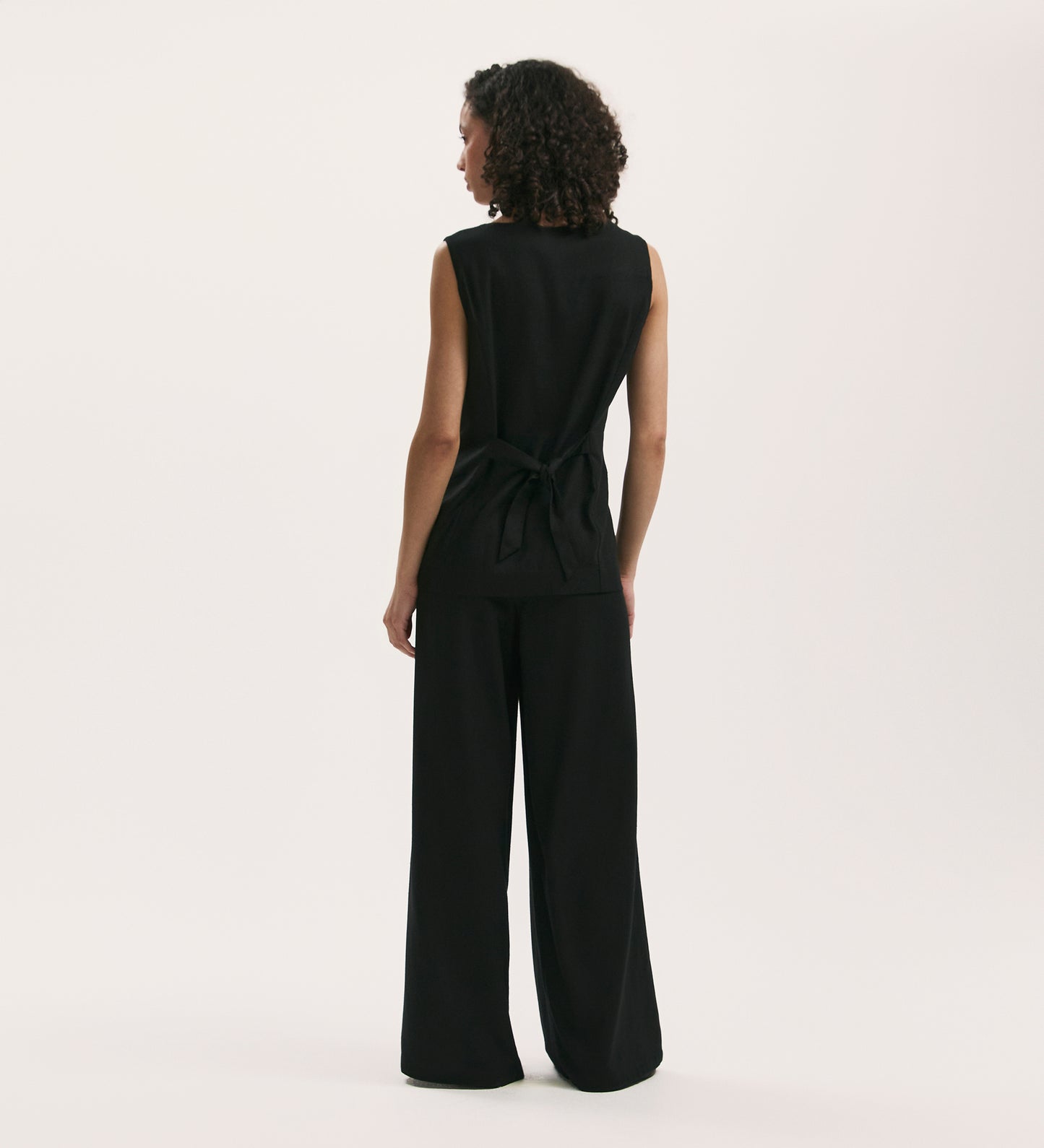 A person with curly hair, seen from behind, is wearing the Vanetia Black Linen Blend Waistcoat with an adjustable tie-back and matching wide-leg pants, standing against a plain white background.