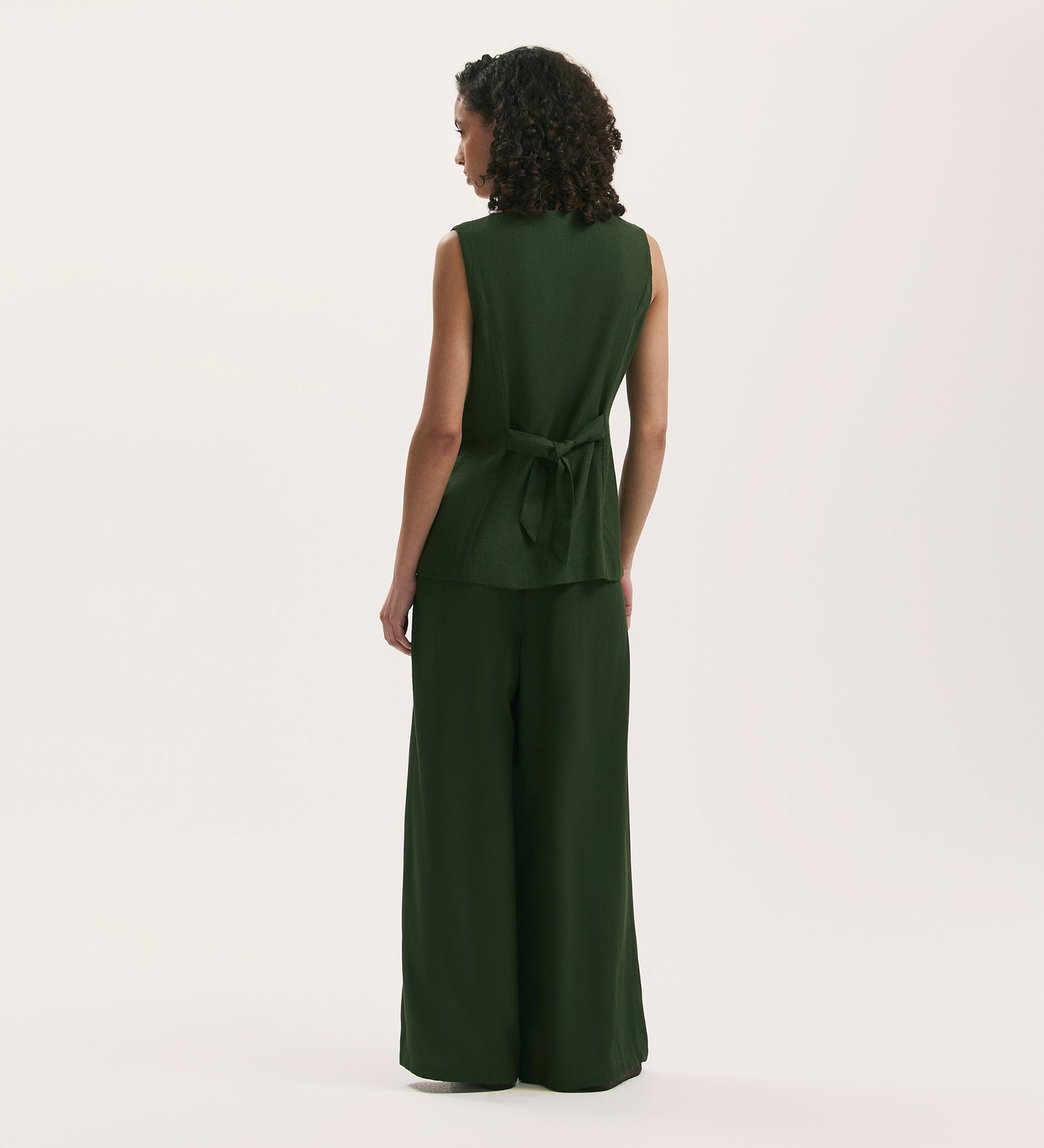 A person with curly hair, seen from behind, is wearing the Vanetia Dark Khaki Linen Blend Waistcoat with matching wide-leg pants, featuring an adjustable tie-back, and standing against a plain light background.