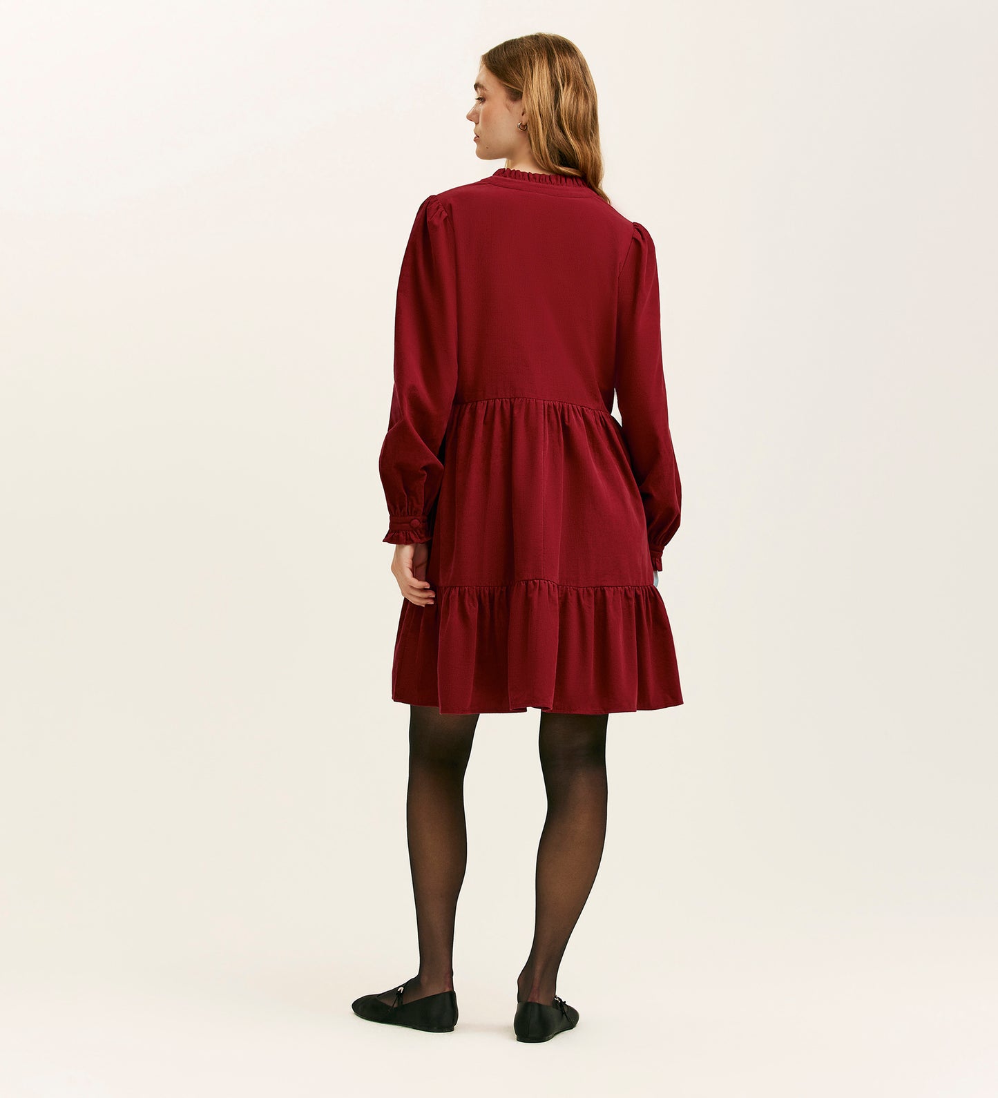 A woman stands facing away, wearing the Verona Rust Corduroy Short Dress with black tights and black shoes against a plain light background.