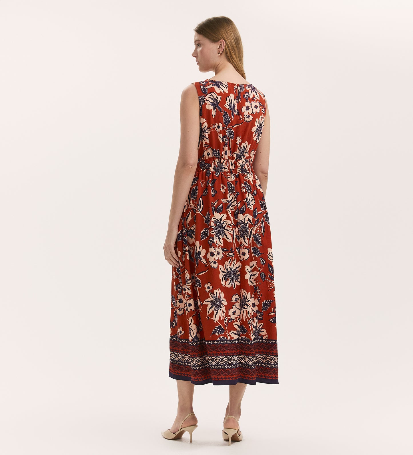 A woman with straight blond hair and light skin faces away, wearing the Willow Rust Floral Sleeveless Midi Dress. The comfortable shirred waist flatters her figure, and she pairs it with beige heeled sandals.