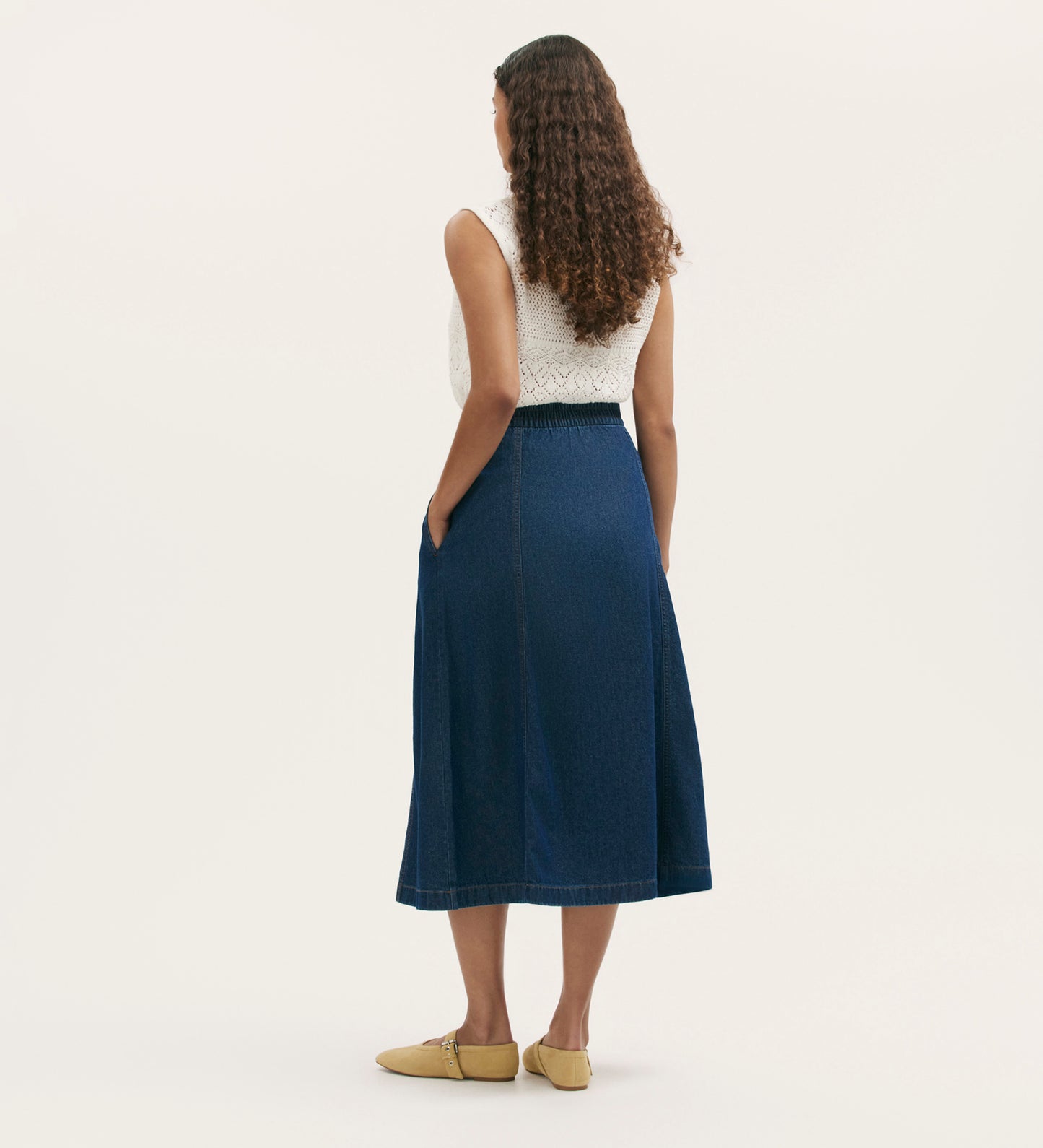 A woman with long curly hair stands facing away in a white sleeveless top, yellow shoes, and the Winona Indigo Denim Skirt—an elastic-waist midi with pockets—set against a plain light background, ideal for casual wardrobes.