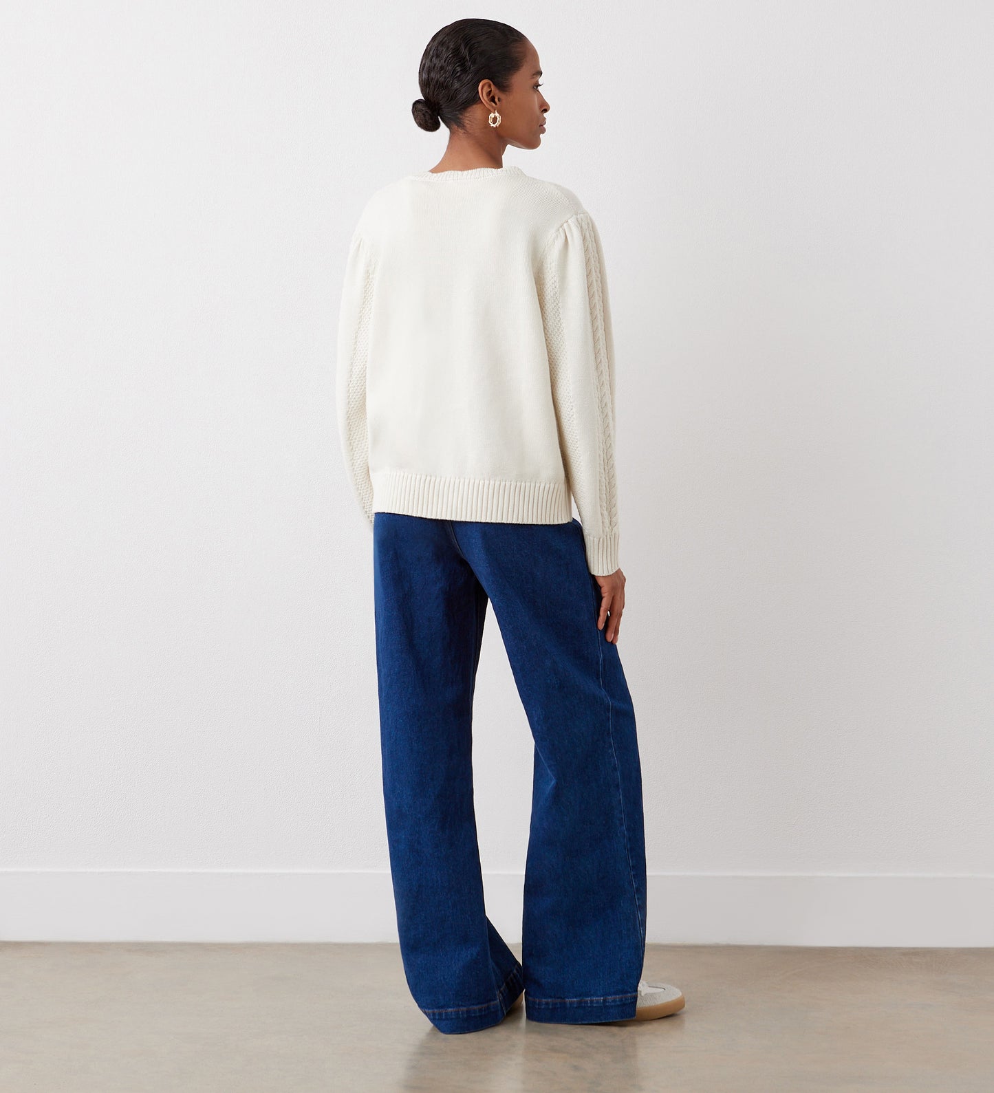 A woman stands facing away, wearing the Sadie Ivory Button Front Cardigan and wide-leg blue jeans. Her hair is tied back, and she is on a light-colored floor against a plain white wall.