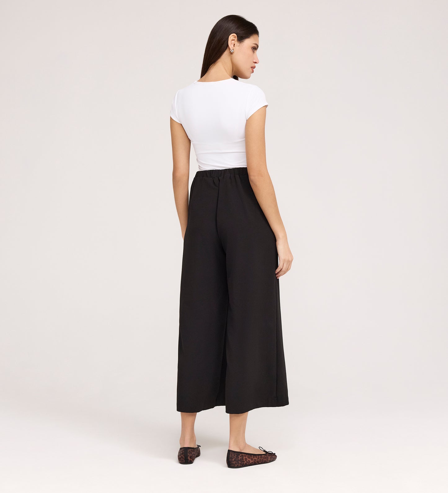 A woman stands against a plain background, facing away. She wears a fitted white short-sleeve top and Simone Black Crepe Wide Leg Trousers with an elastic waistband, dark flat shoes, and her straight hair loose at her sides.