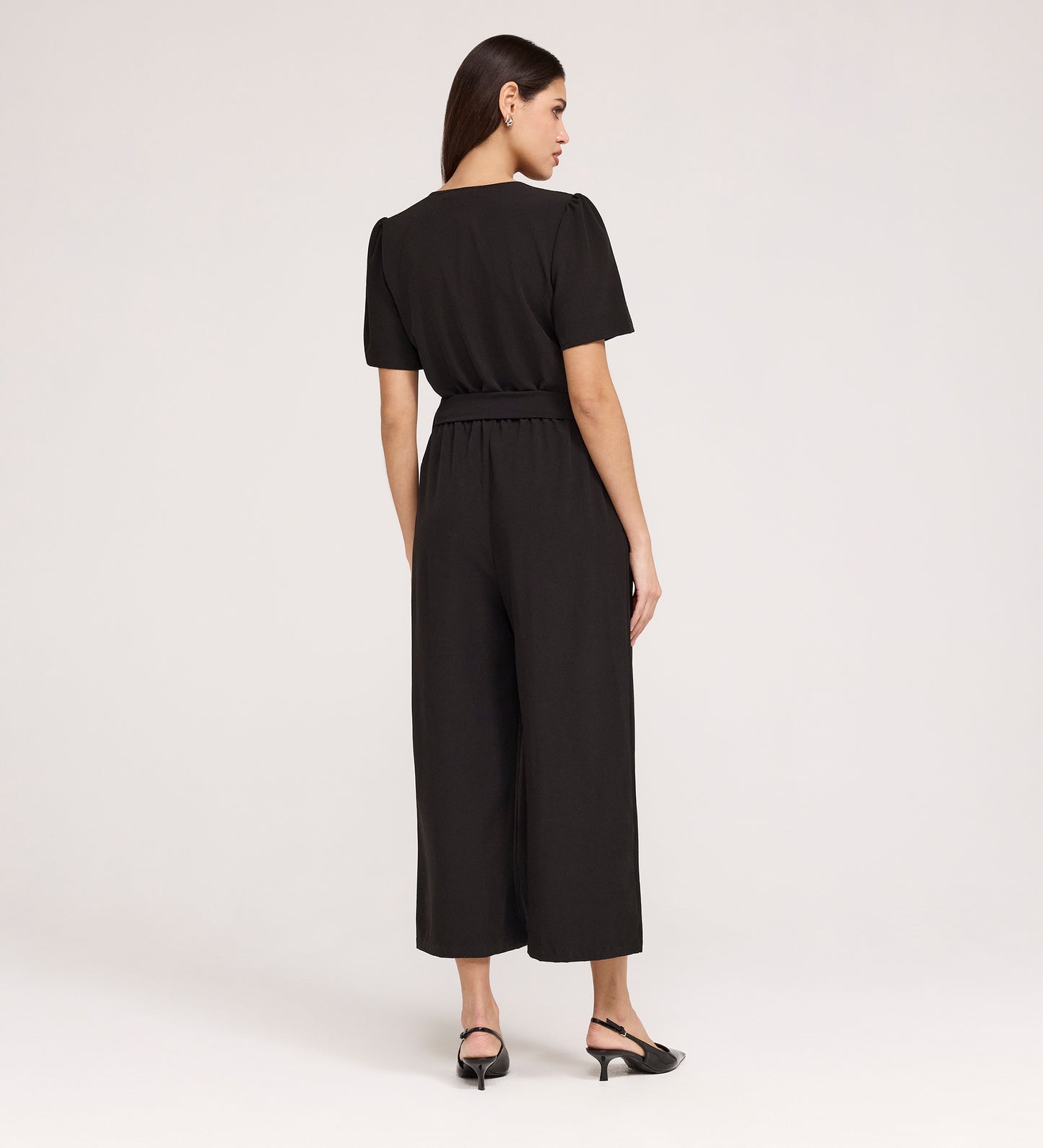 A woman, facing away, models the Uma Black Crepe Short Sleeve Jumpsuit featuring a tie waist and cropped wide-leg hems. She pairs it with black heeled sandals. Her straight dark hair complements the simple, light background.