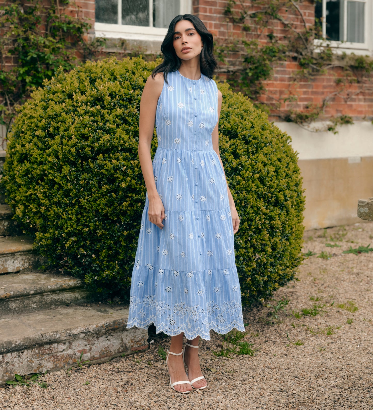 Woman in Rose Blue Stripe Cotton Broderie Sleeveless Midi Dress standing outdoors with greenery and a brick building in the background