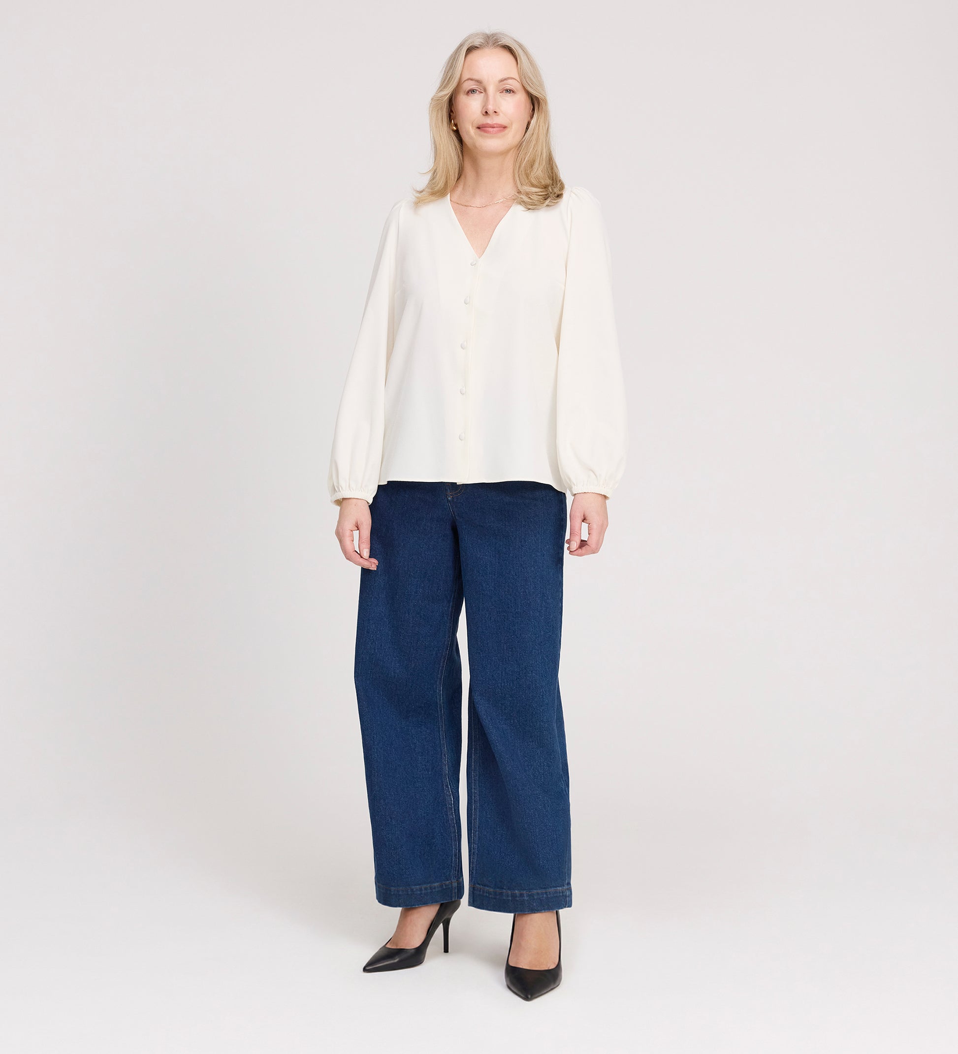 A woman with long blonde hair stands against a light background, wearing the Elvora Ivory Button Up V-neck Top, paired with wide-leg blue jeans and black high-heeled shoes. She is smiling slightly and facing the camera.