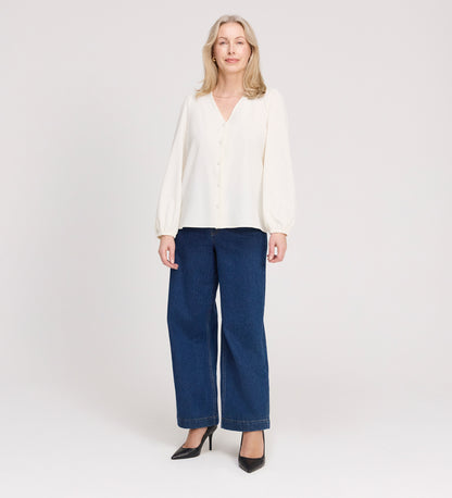 A woman with long blonde hair stands against a light background, wearing the Elvora Ivory Button Up V-neck Top, paired with wide-leg blue jeans and black high-heeled shoes. She is smiling slightly and facing the camera.