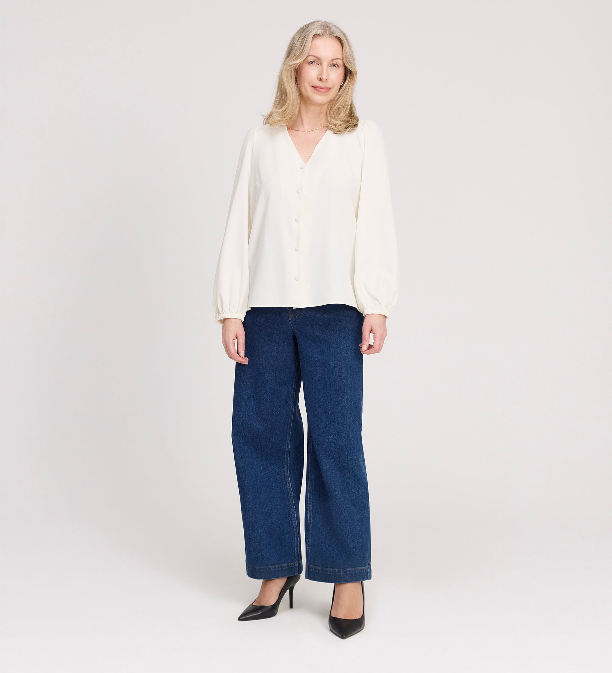 A woman with long, light blonde hair is standing and smiling, wearing the Elvora Ivory Button Up V-neck Top, wide-leg blue jeans, and black high heels against a plain light background.