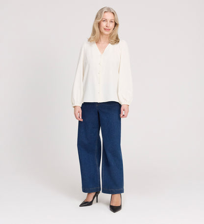 A woman with long, light blonde hair is standing and smiling, wearing the Elvora Ivory Button Up V-neck Top, wide-leg blue jeans, and black high heels against a plain light background.