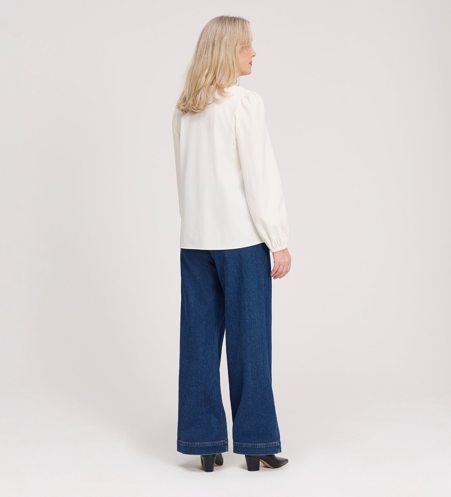 A woman with blonde hair wears the Elvora Ivory Button Up V-neck Top, paired with long sleeves, wide-leg blue jeans, and black heels, standing and facing away from the camera against a plain light background.