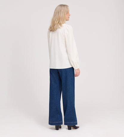 A woman with blonde hair wears the Elvora Ivory Button Up V-neck Top, paired with long sleeves, wide-leg blue jeans, and black heels, standing and facing away from the camera against a plain light background.