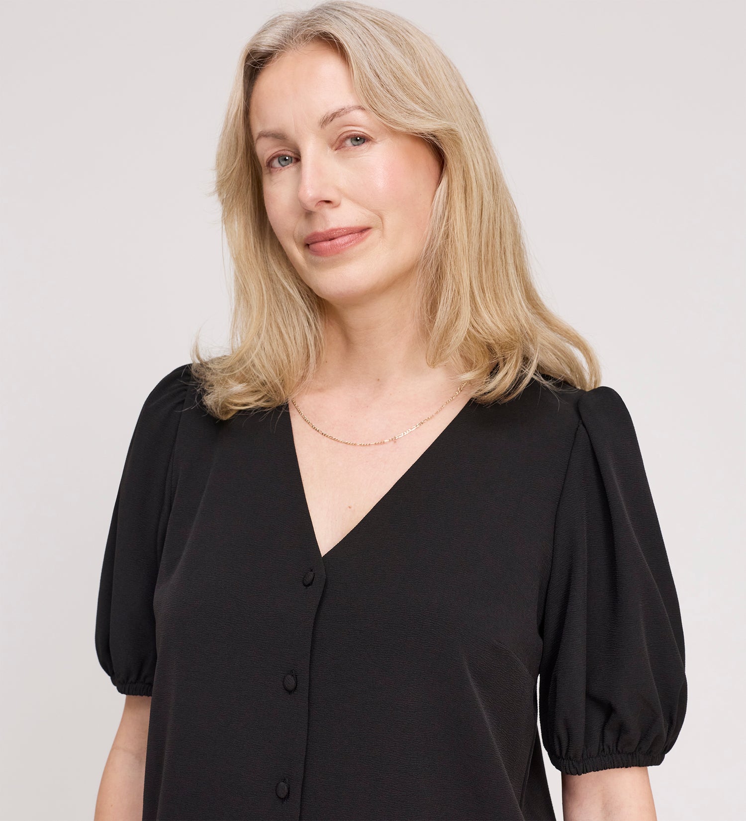 A woman with light blonde hair wears the Fleur Black Button Down Short Sleeve Top and a delicate necklace, smiling softly against a plain light background.