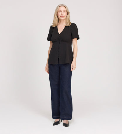 A woman with light hair stands against a plain background, wearing the Maryam Black Button Up V-Neck Short Sleeve Top, dark blue wide-leg jeans, and black heels. She smiles slightly with her arms relaxed by her sides.