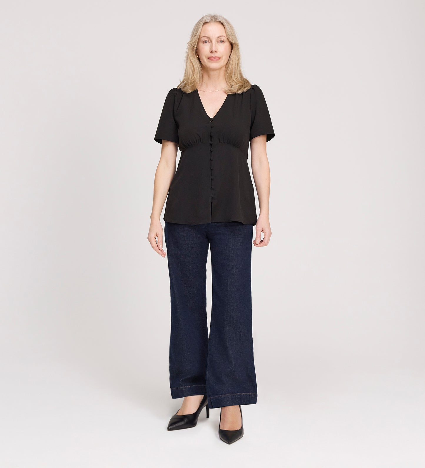 A woman with light hair faces forward, wearing a Maryam Black Button Up V-Neck Short Sleeve Top, paired with dark wide-leg jeans and black heels, against a plain light background.