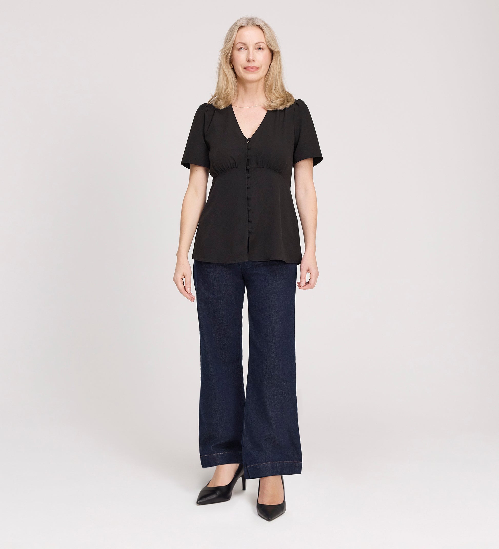 A woman with light hair faces forward, wearing a Maryam Black Button Up V-Neck Short Sleeve Top, paired with dark wide-leg jeans and black heels, against a plain light background.