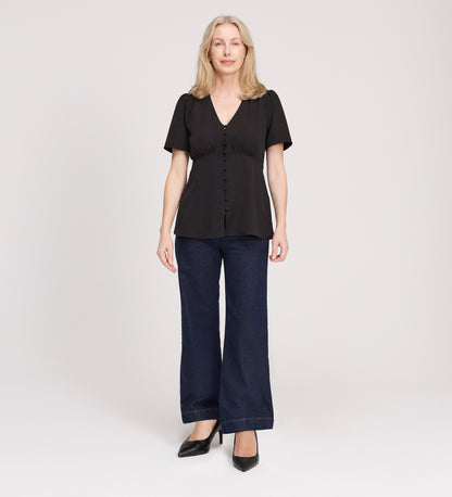 A woman with light hair faces forward, wearing a Maryam Black Button Up V-Neck Short Sleeve Top, paired with dark wide-leg jeans and black heels, against a plain light background.