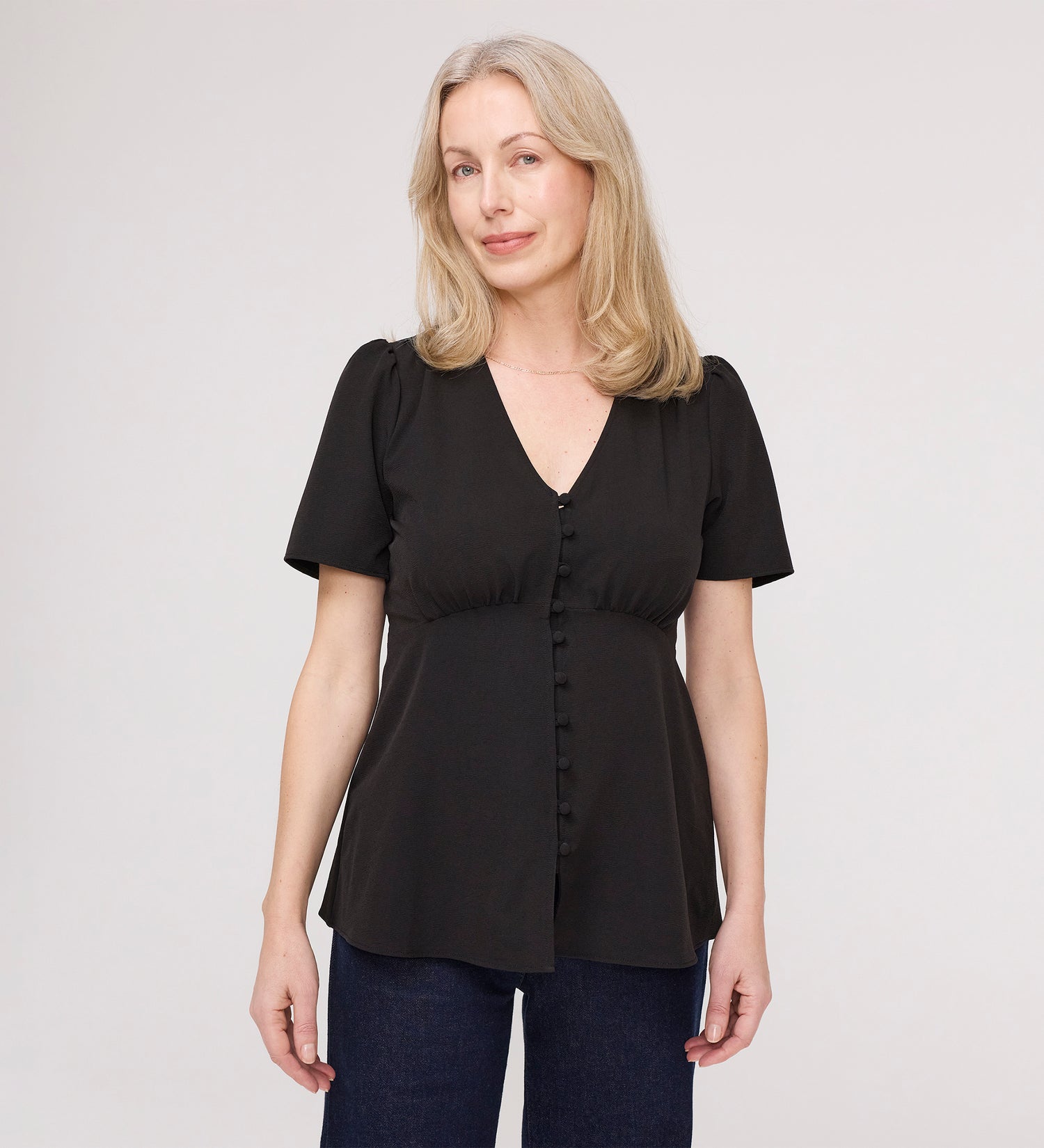 A woman with light blonde hair wears the Maryam Black Button Up V-Neck Short Sleeve Top, standing against a plain light background and looking at the camera with a slight smile.