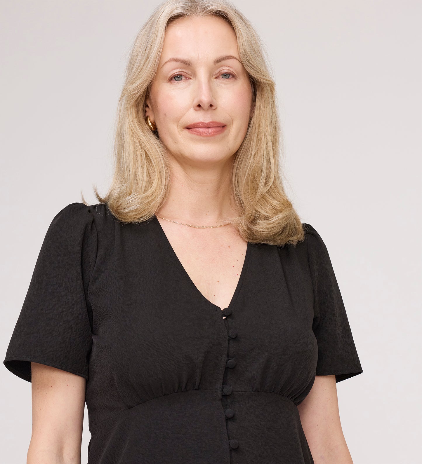 A woman with shoulder-length blonde hair wears the Maryam Black Button Up V-Neck Short Sleeve Top, standing against a plain light background and looking at the camera with a neutral expression.